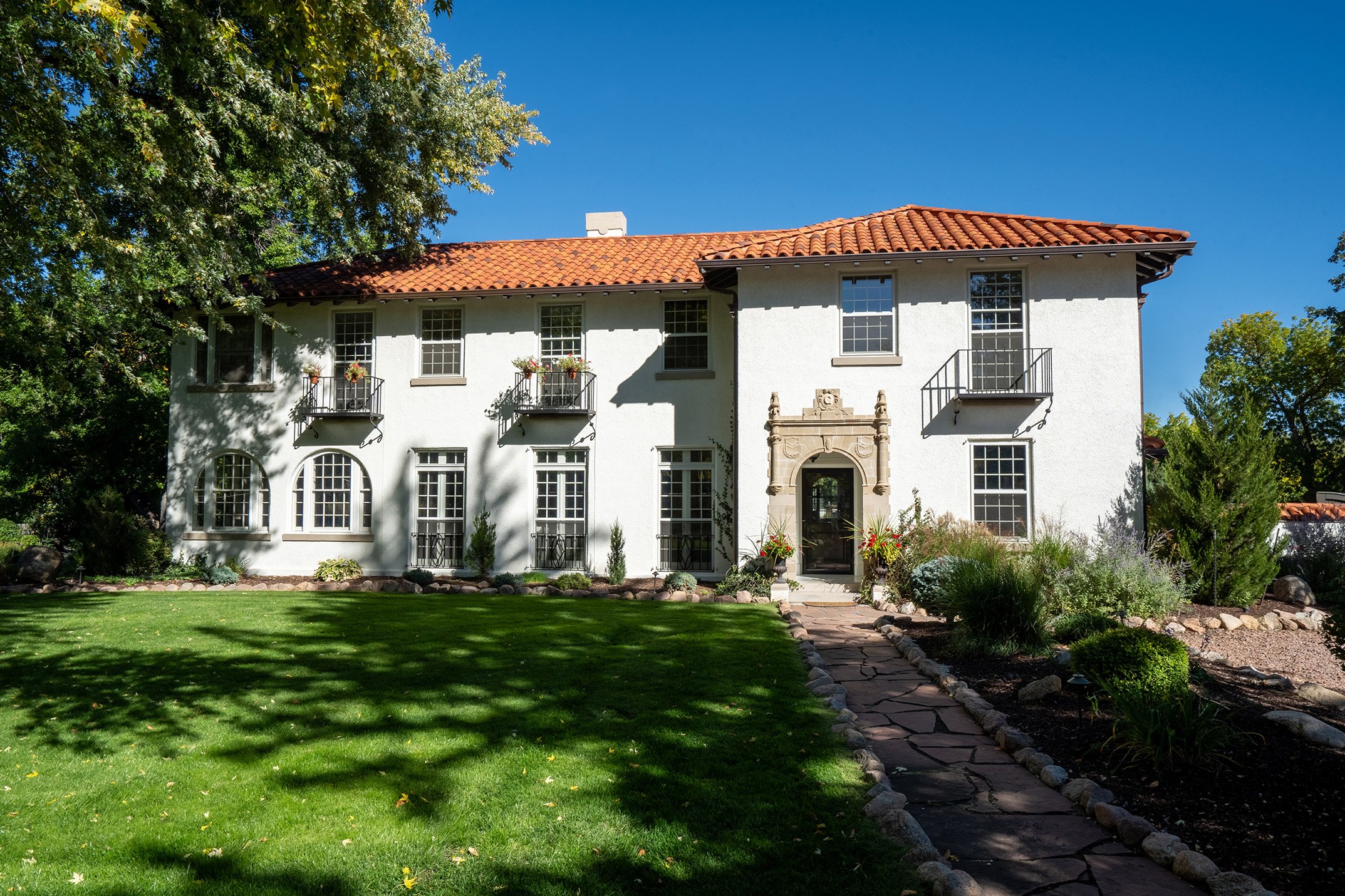 Spanish-style stucco house with terracotta tile roof, arched windows, landscaped garden, and stone walkway under a clear blue sky