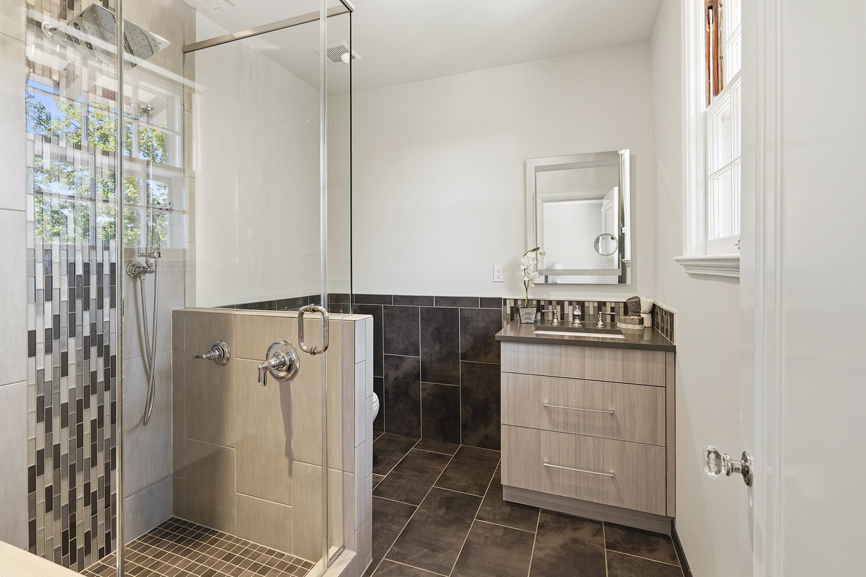 Modern bathroom with walk-in shower, dark tiled floors, light wood vanity, white walls, and a large window.
