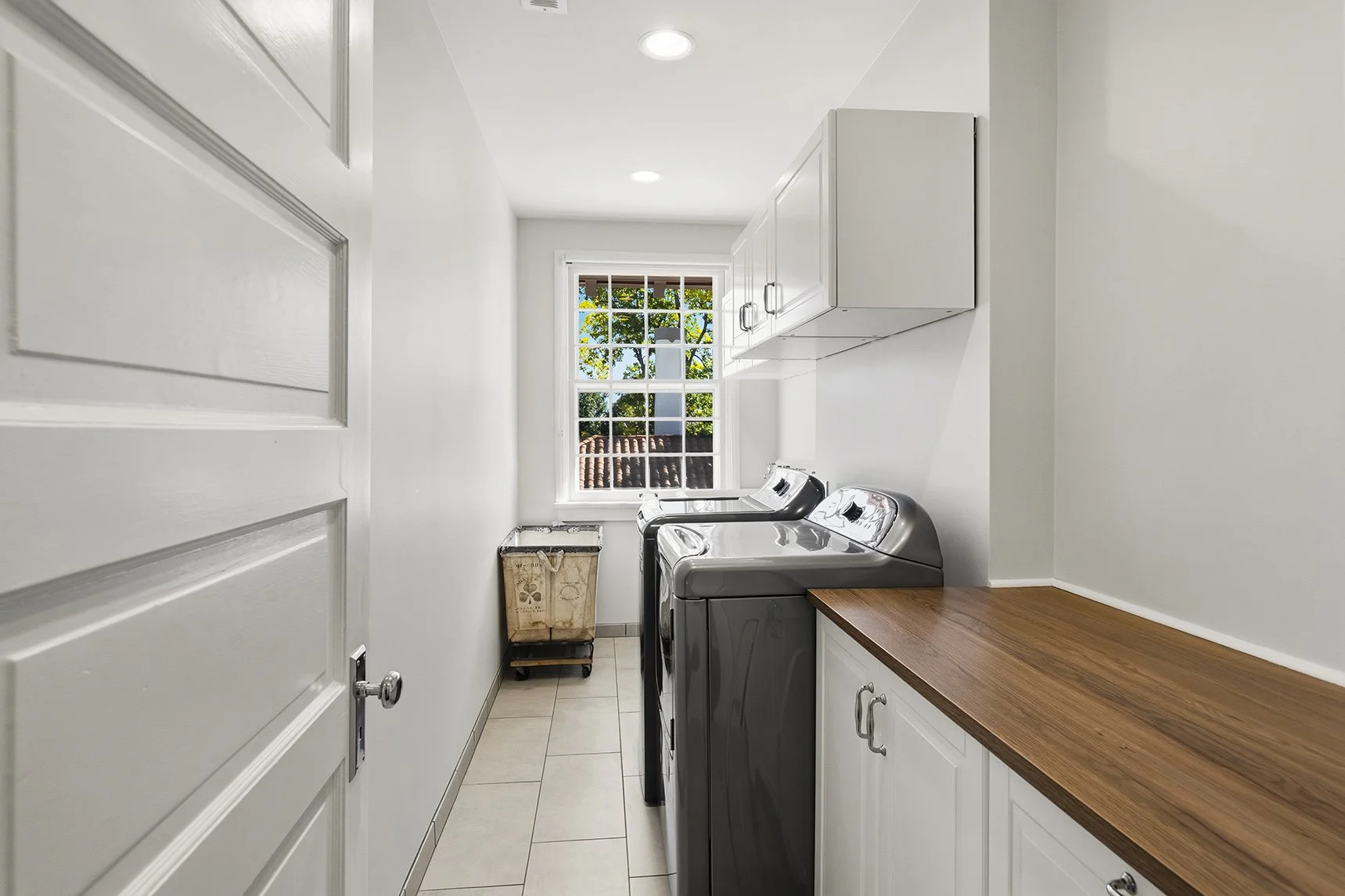 Bright laundry room with front-loading washer and dryer, white cabinets, wood countertop, tiled floor, and window overlooking trees