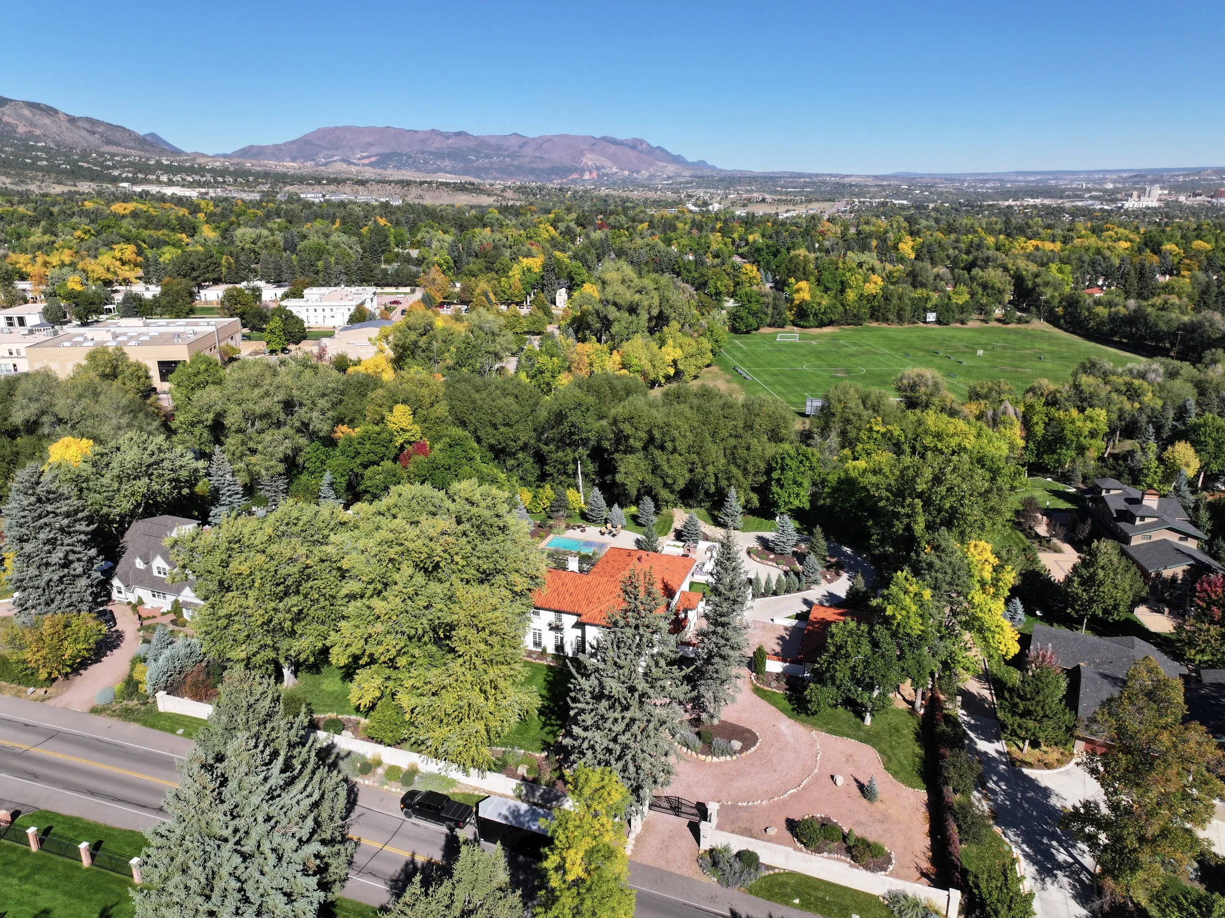 Aerial view of a leafy residential neighborhood with autumn trees, soccer field, and mountain range under a clear blue sky