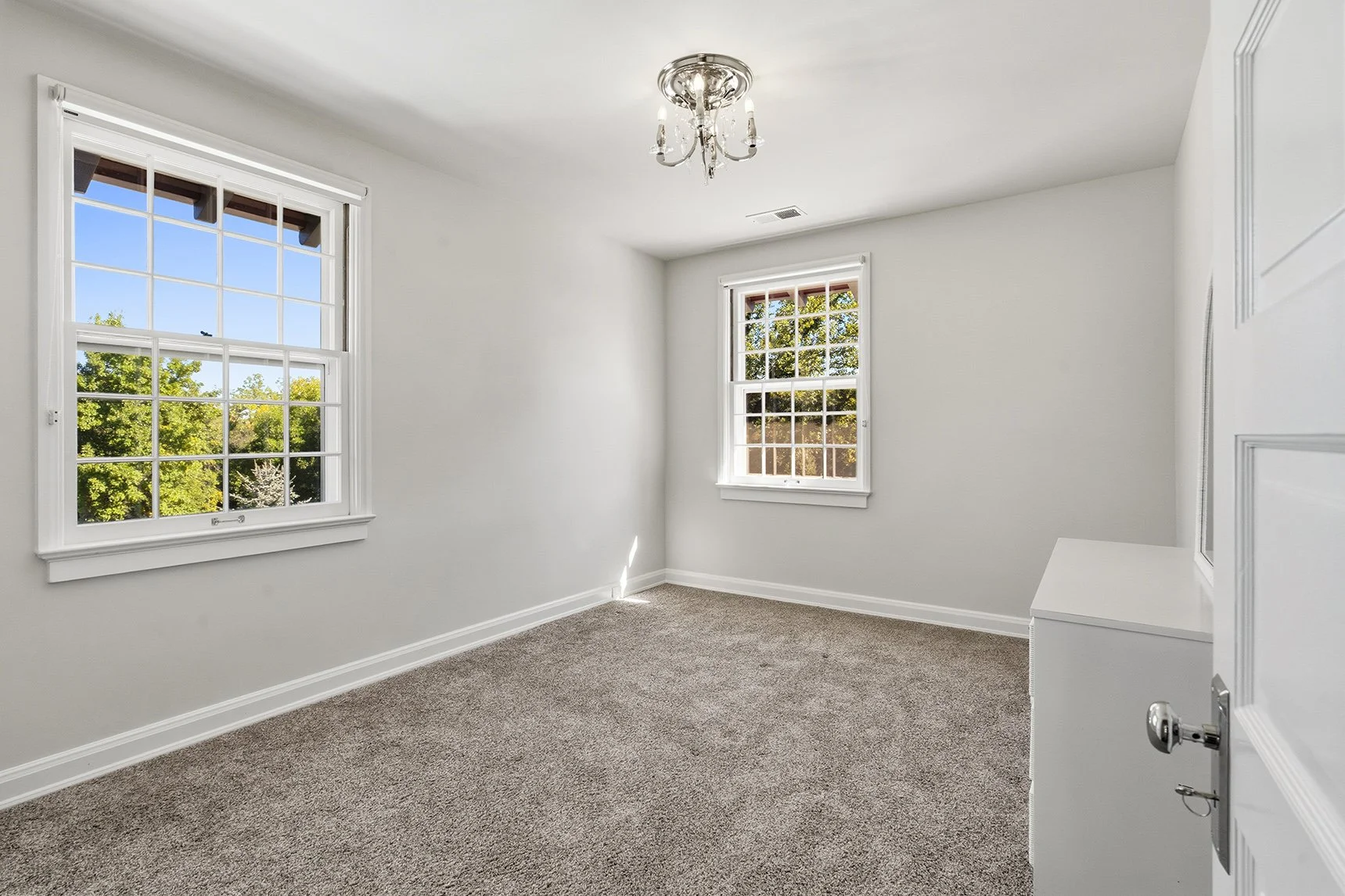 Bright empty bedroom with gray carpet, white walls, two large windows, and a crystal chandelier