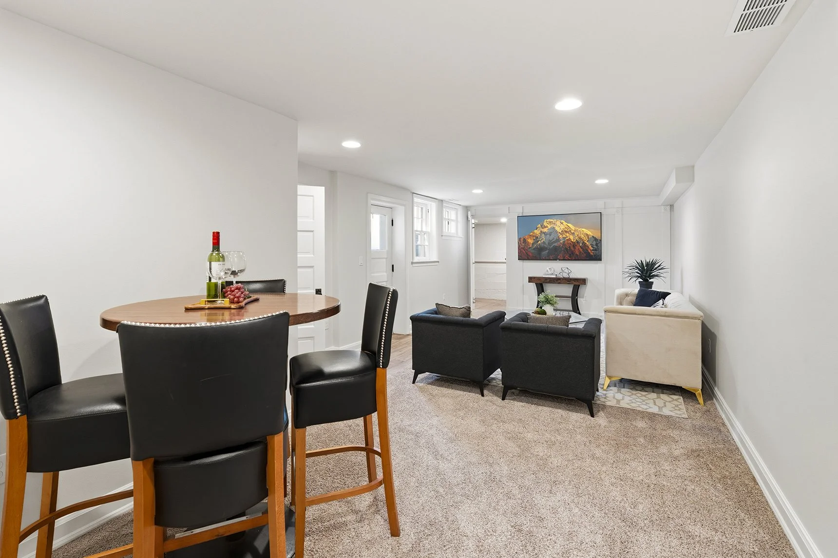Bright finished basement with a dining table, black bar stools, wine, and a furnished living area with a wall-mounted mountain landscape TV