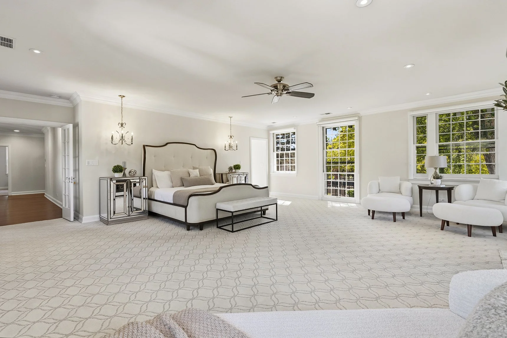 Spacious elegant bedroom with upholstered bed, chandeliers, ceiling fan, seating area, large windows, and patterned carpet