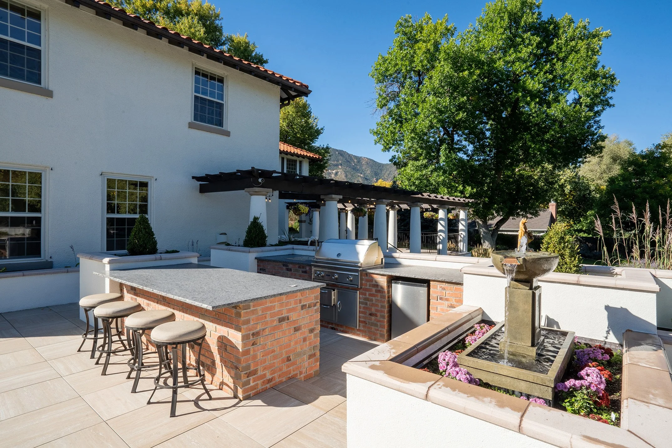 Luxury outdoor kitchen with brick island, grill, bar stools, pergola, fountain, and mountain views