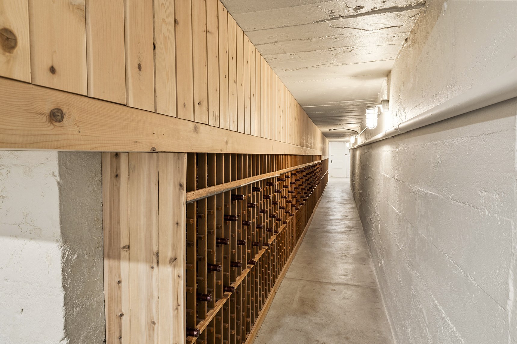 A narrow wine cellar corridor with a long wooden wine rack on the left side and white concrete walls on the right, with fluorescent lights.