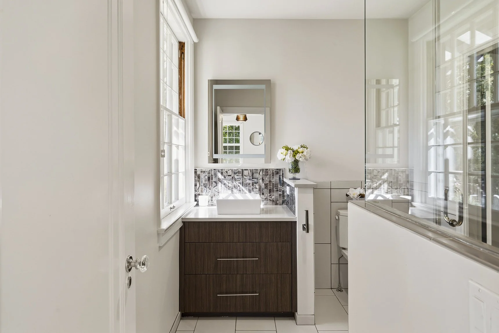 Bright modern bathroom with dark wood vanity, white countertop, patterned tile backsplash, large mirror, windows, bathtub, and flower arrangement