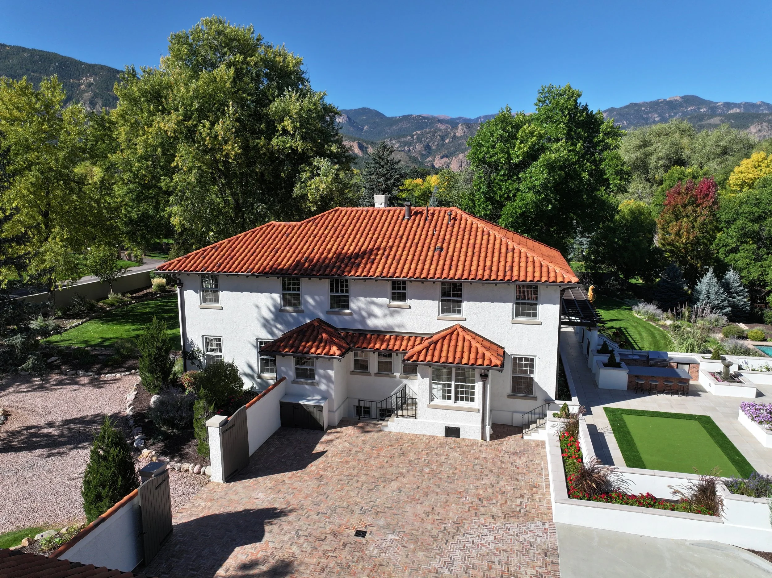 A Spanish-style stucco house with red tile roofs, landscaped courtyard, putting green, and mountain views under a clear blue sky.