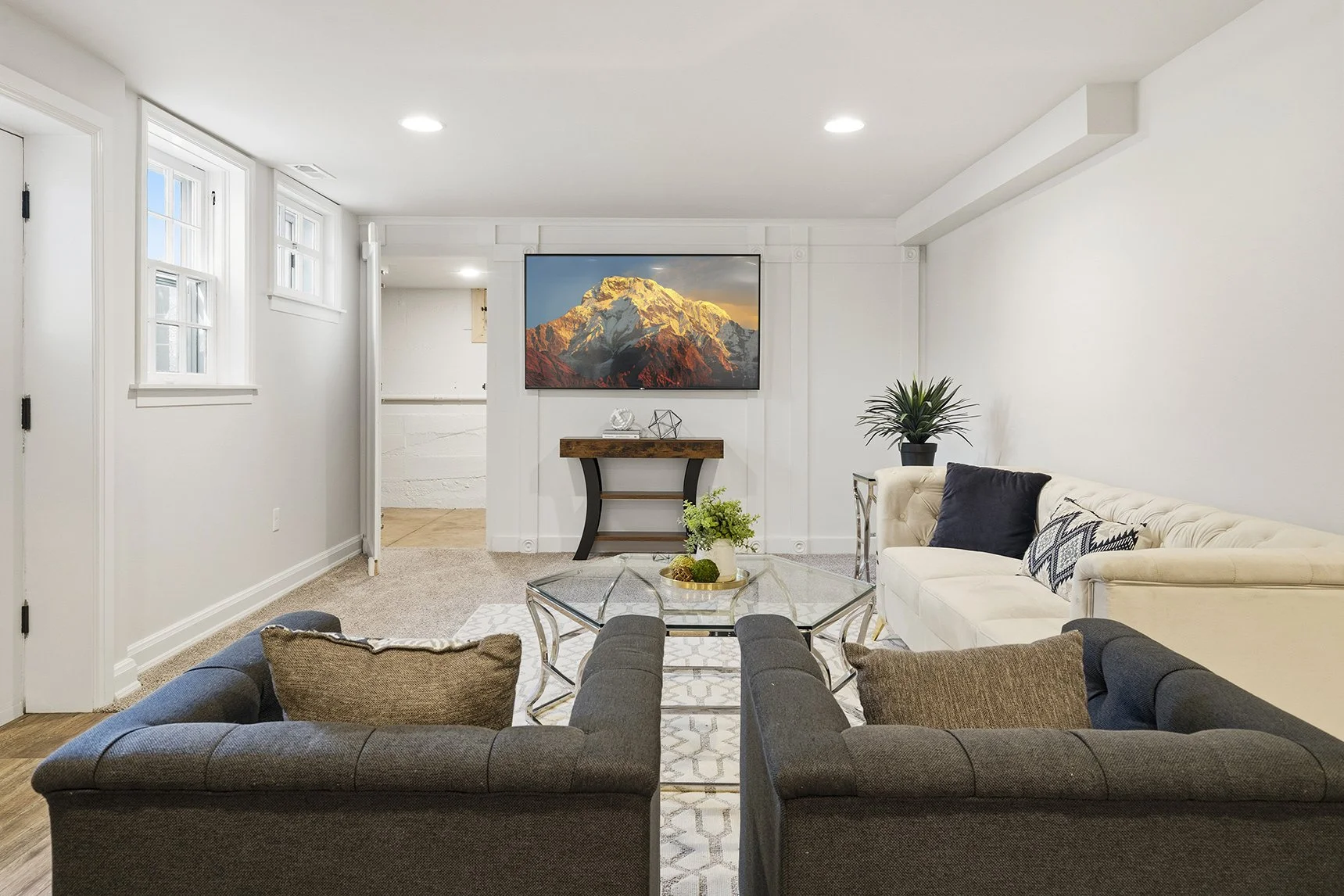 Bright modern living room with sofas, glass coffee table, wall-mounted TV, mountain landscape artwork, console table, and potted plants