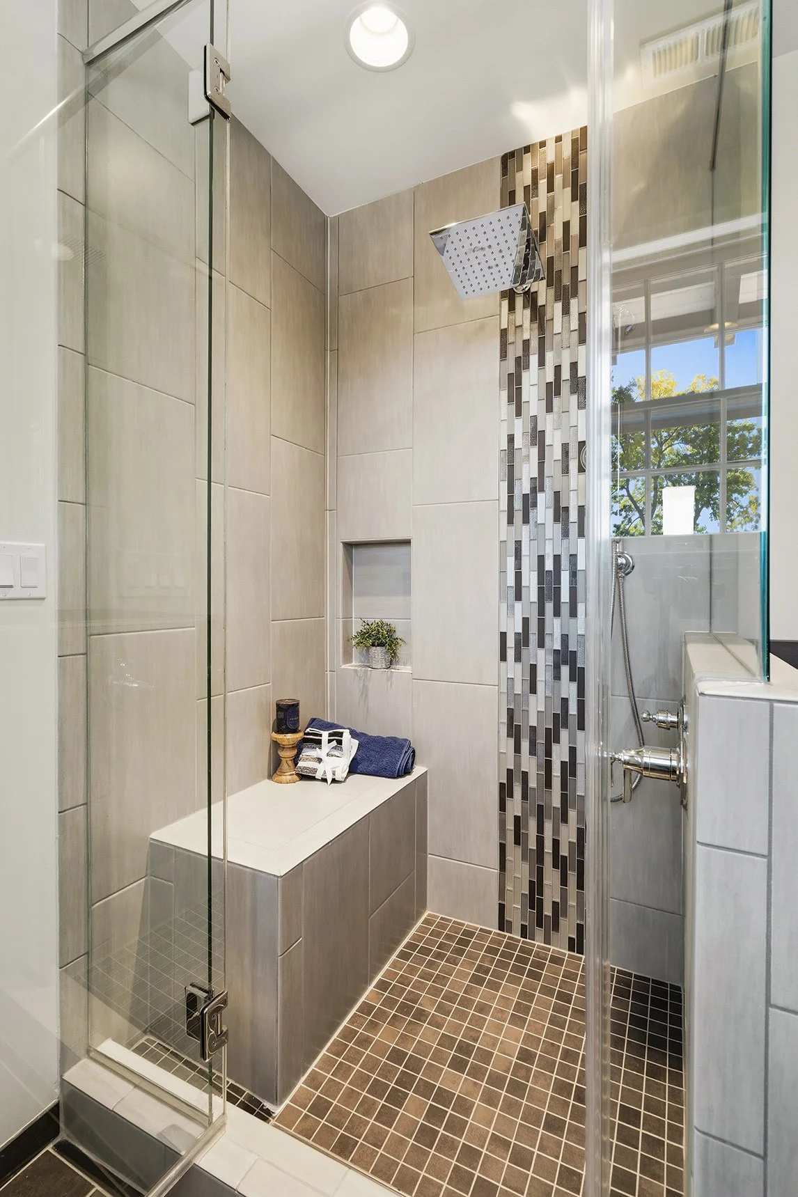 Modern shower with beige tiles, a vertical strip of decorative mosaic tiles, a built-in bench with towels and toiletries, a small shelf with a plant, and a glass door with a metal handle. A window shows trees outside.