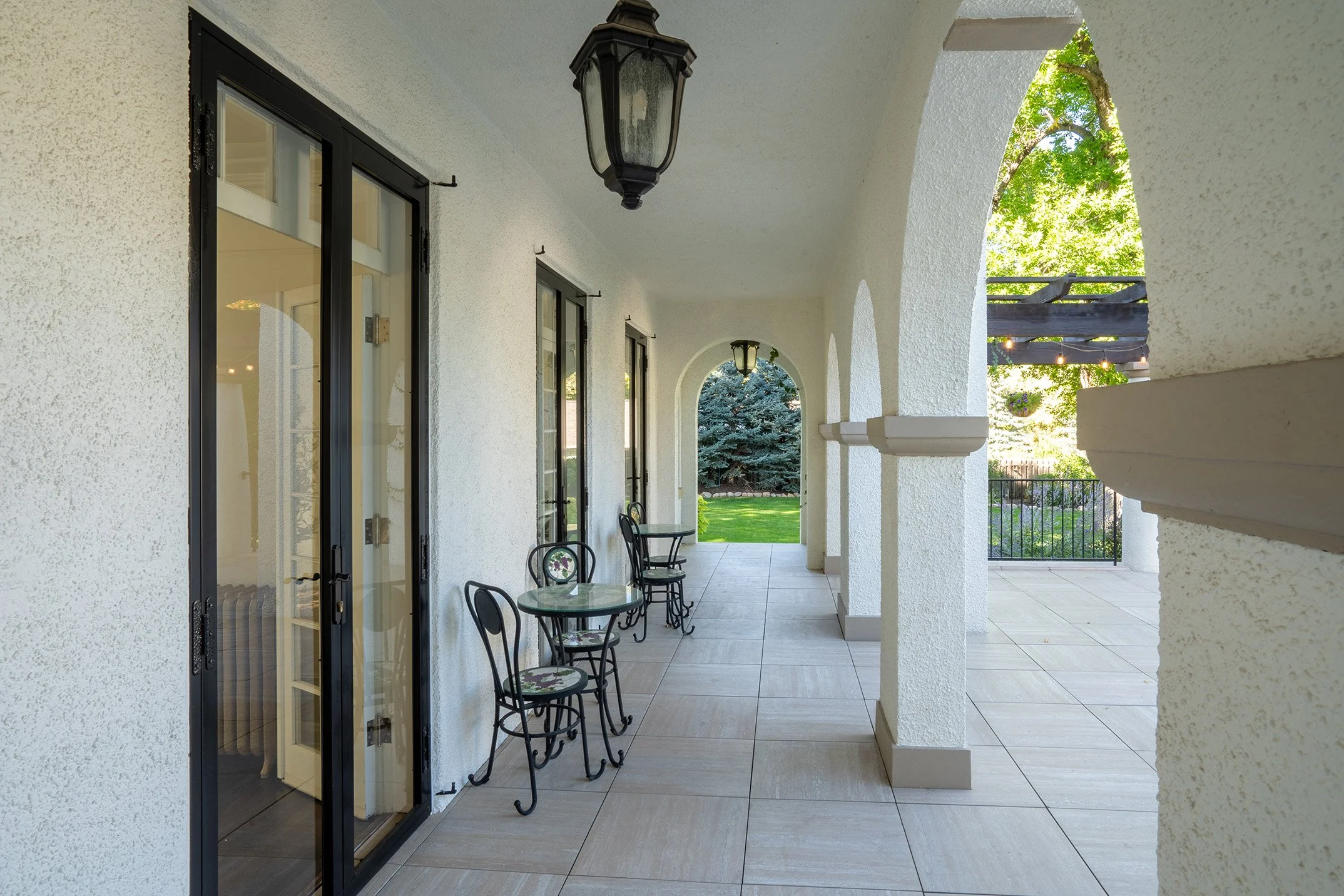 Covered porch with white stucco walls, black framed glass door, black chairs, and a view of green lawn and trees.