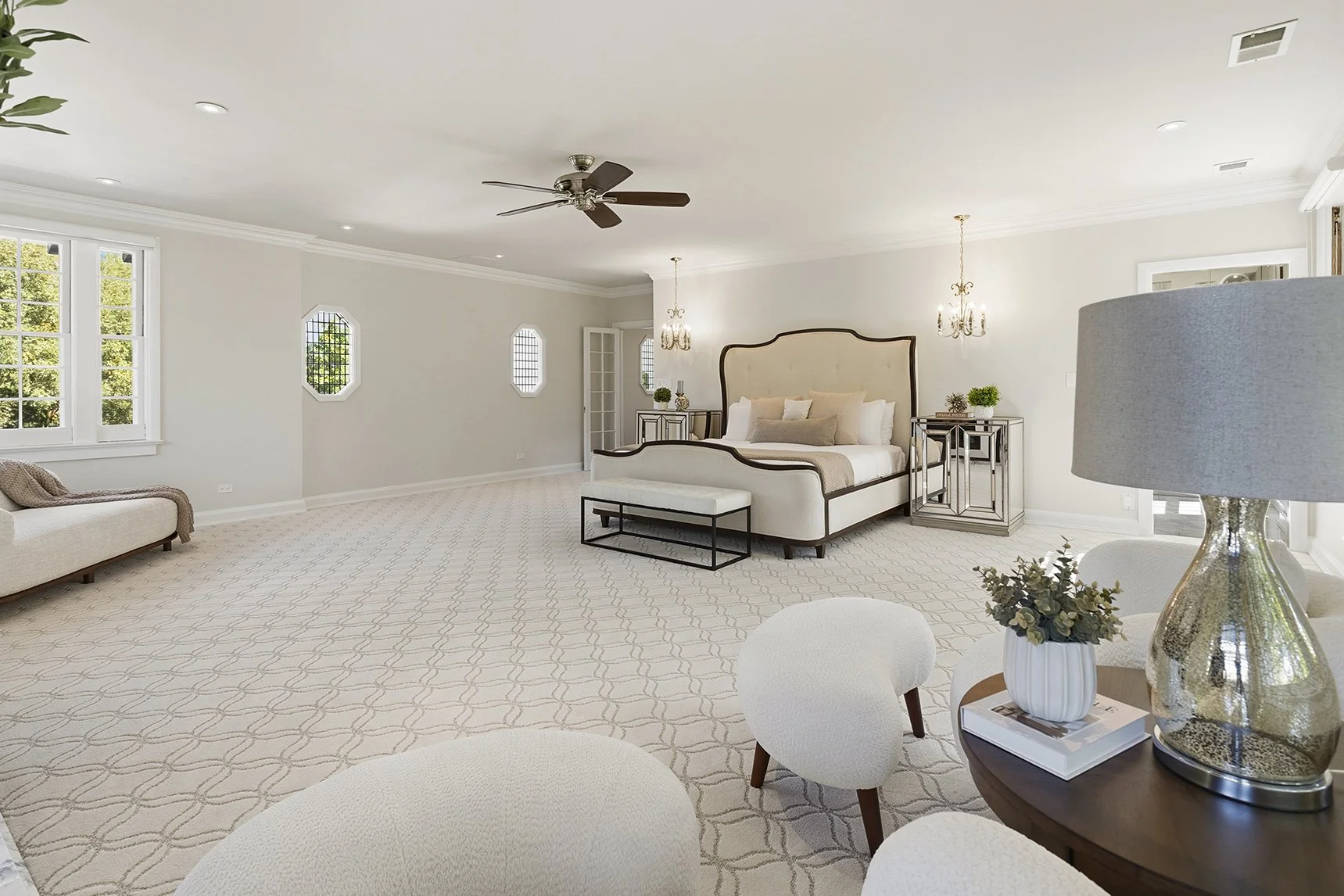 Spacious elegant bedroom with upholstered bed, chandeliers, ceiling fan, patterned carpet, and sitting area
