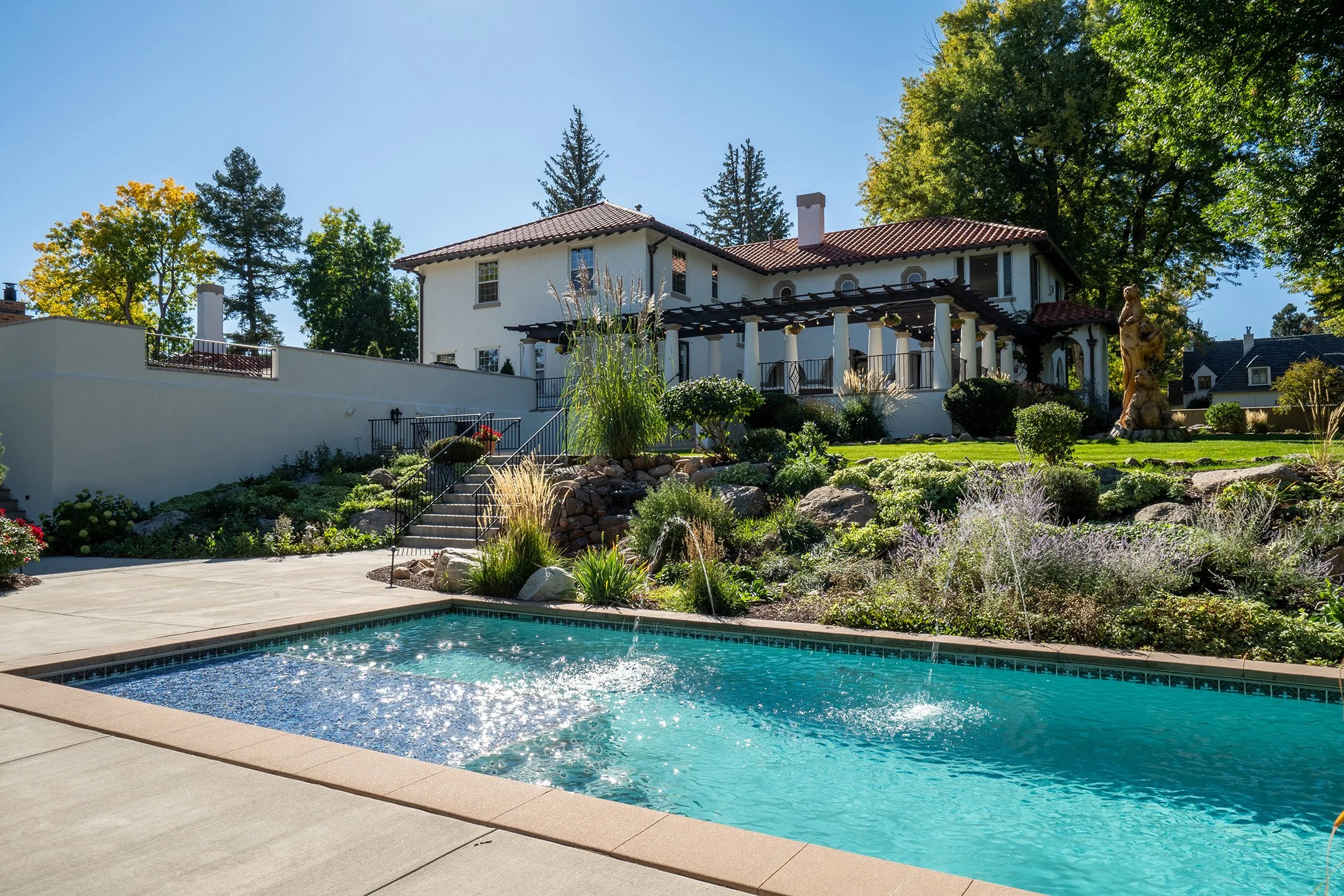 A backyard with a swimming pool, surrounded by a concrete deck, leading to a landscaped yard with rocks and plants, and a large house with a red-tiled roof and white walls in the background, along with tall trees and a clear blue sky.