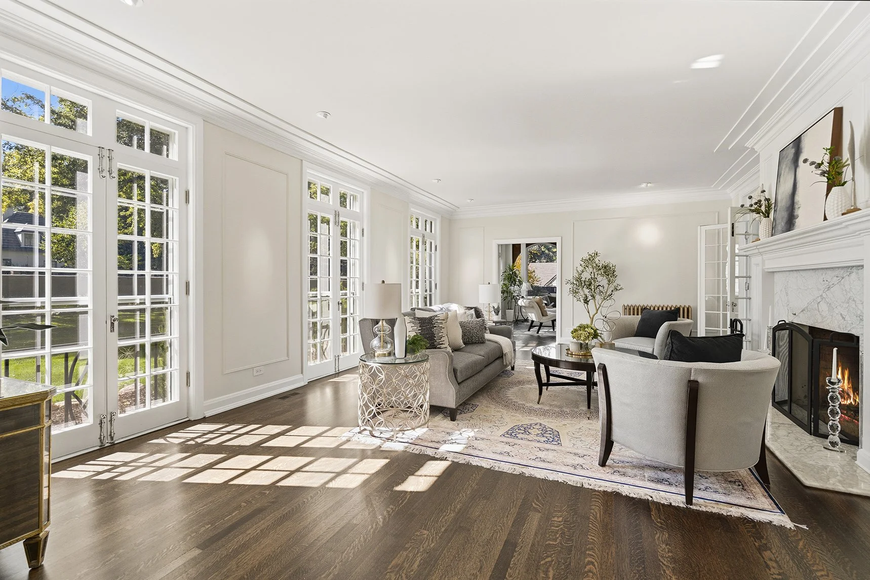Bright living room with white walls, large windows, and hardwood floors, furnished with sofas, armchairs, tables, and a fireplace.