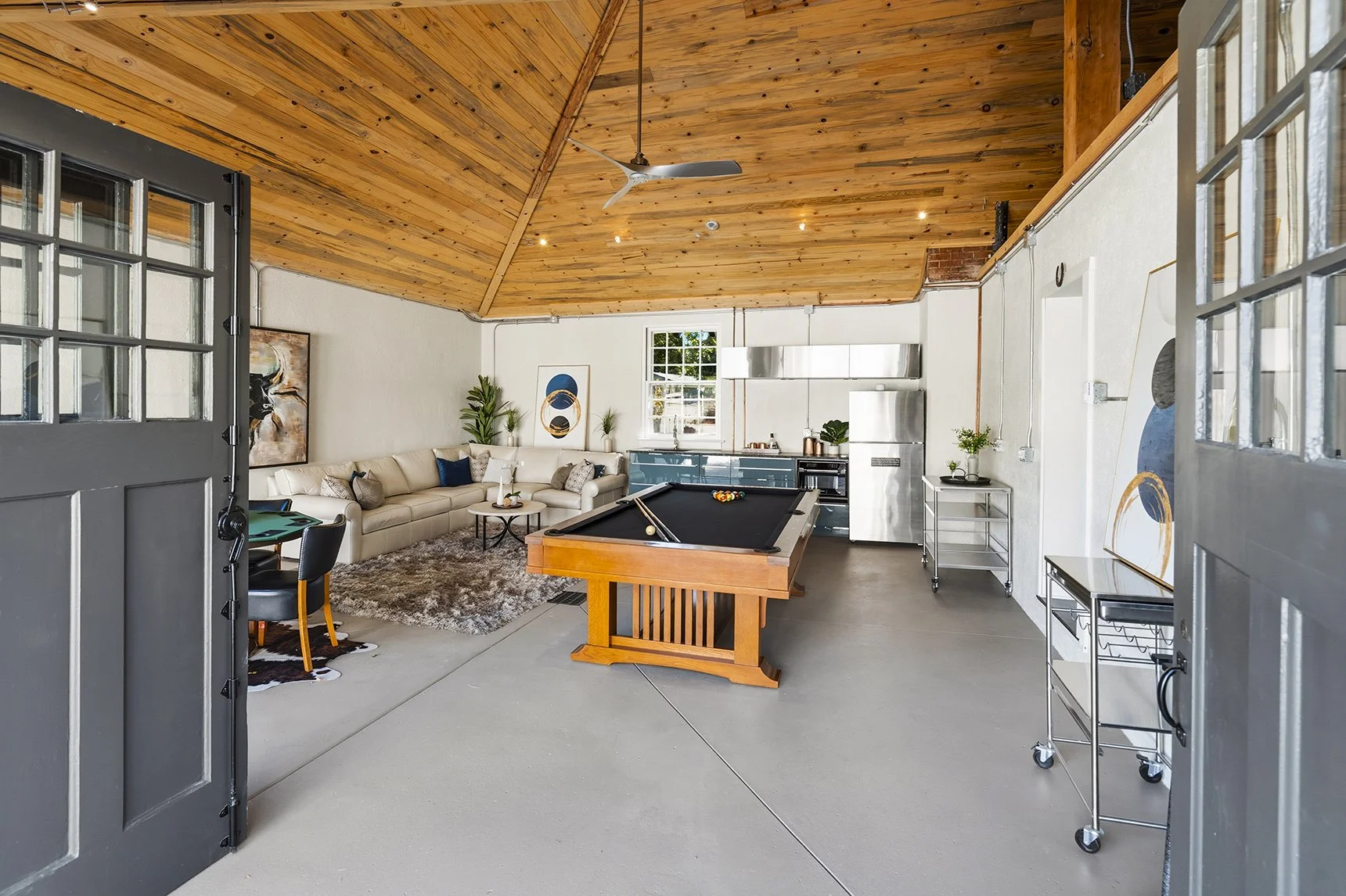 Interior view of a spacious modern room with a high wood-paneled ceiling, a pool table, a beige sectional sofa with decorative pillows, a shaggy rug, a small round coffee table, a blue kitchen with stainless steel appliances, and artwork on the walls