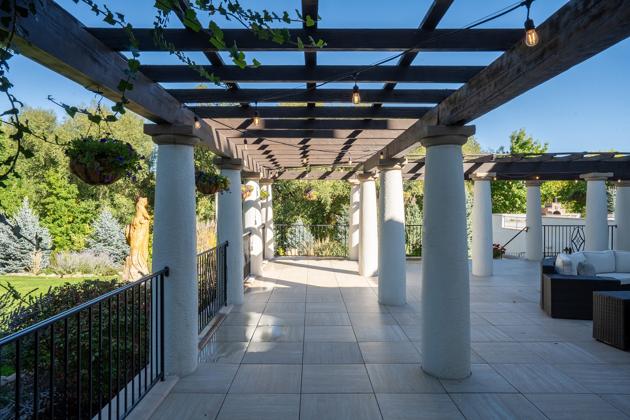 A covered outdoor patio with a wooden pergola supported by white round columns. String lights hang from the pergola. The patio has light-colored tiled flooring and is surrounded by lush green trees and plants, with hanging flower baskets and outdoor 