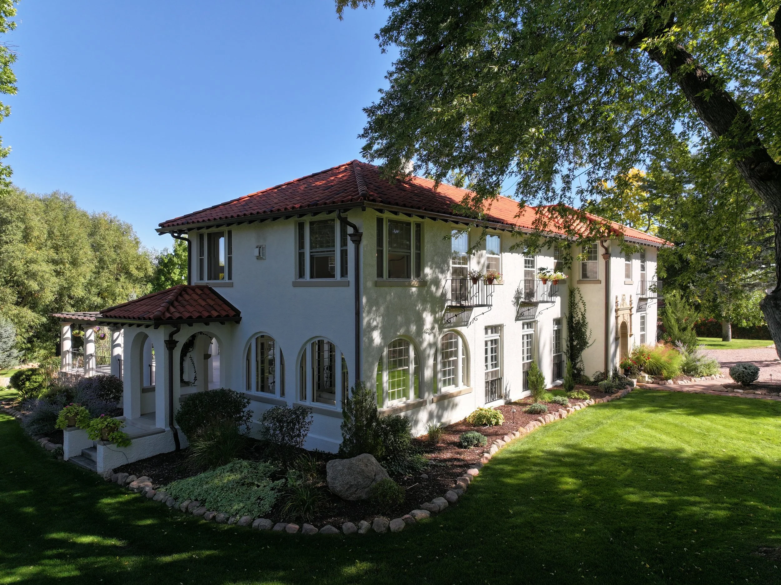 A large white house with a red tile roof, multiple windows, and a front porch with archways, surrounded by a green lawn, trees, and a landscaped garden.