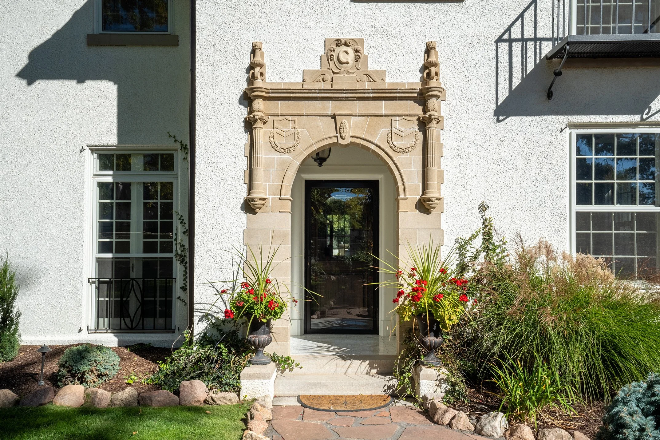 Elegant white stucco home entrance with ornate stone archway, black door, flower planters, and landscaped garden