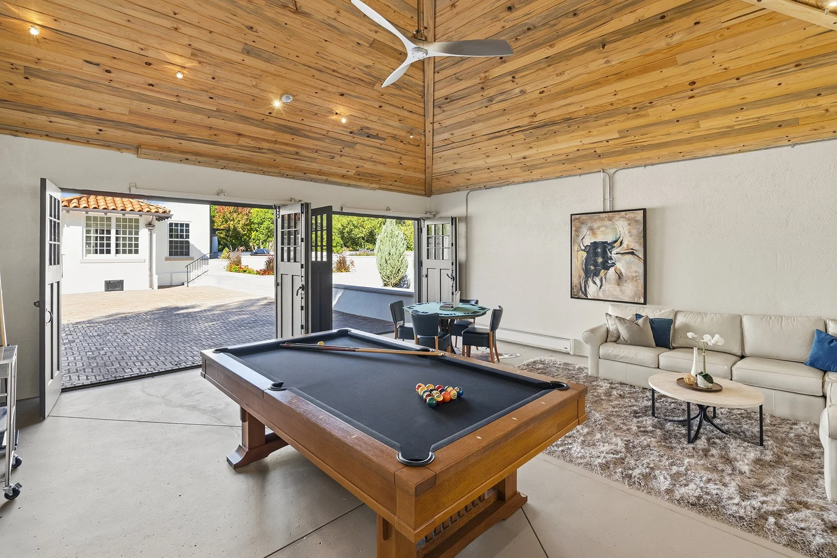 Bright game room with a pool table, seating area, wood-paneled vaulted ceiling, ceiling fan, and open French doors leading to a courtyard