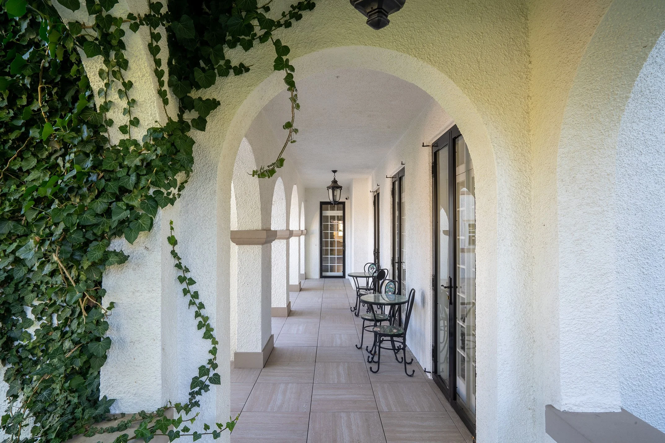 Covered white stucco veranda with arched columns, ivy-covered wall, French doors, lantern lighting, and small café tables and chairs.