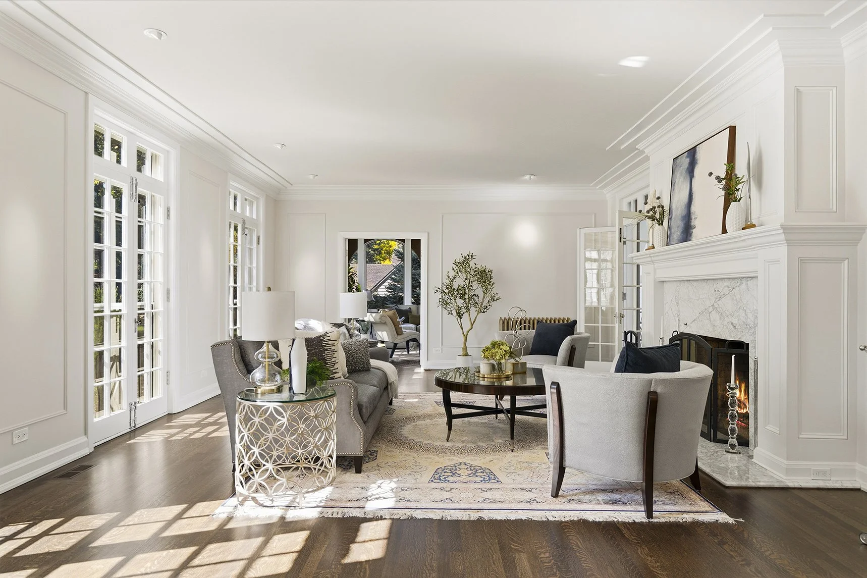 Bright and spacious living room with white walls, large windows, and hardwood floors. Features include a fireplace, gray sofas, armchairs, a round coffee table with decorative objects, and contemporary artwork above the mantel. Natural light casts sh