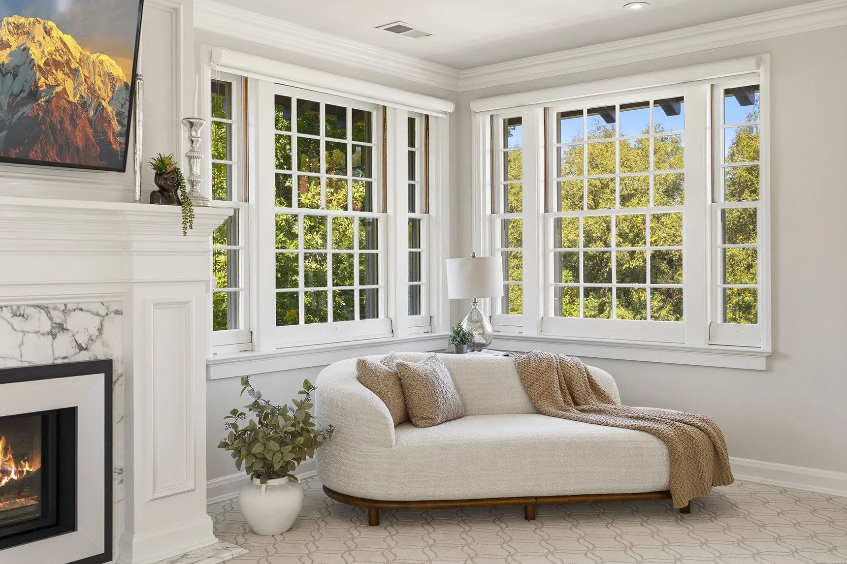 Living room with large windows showing outside green trees, a white couch with pillows and a blanket, a lamp, a potted plant, and a fireplace with a marble surround.
