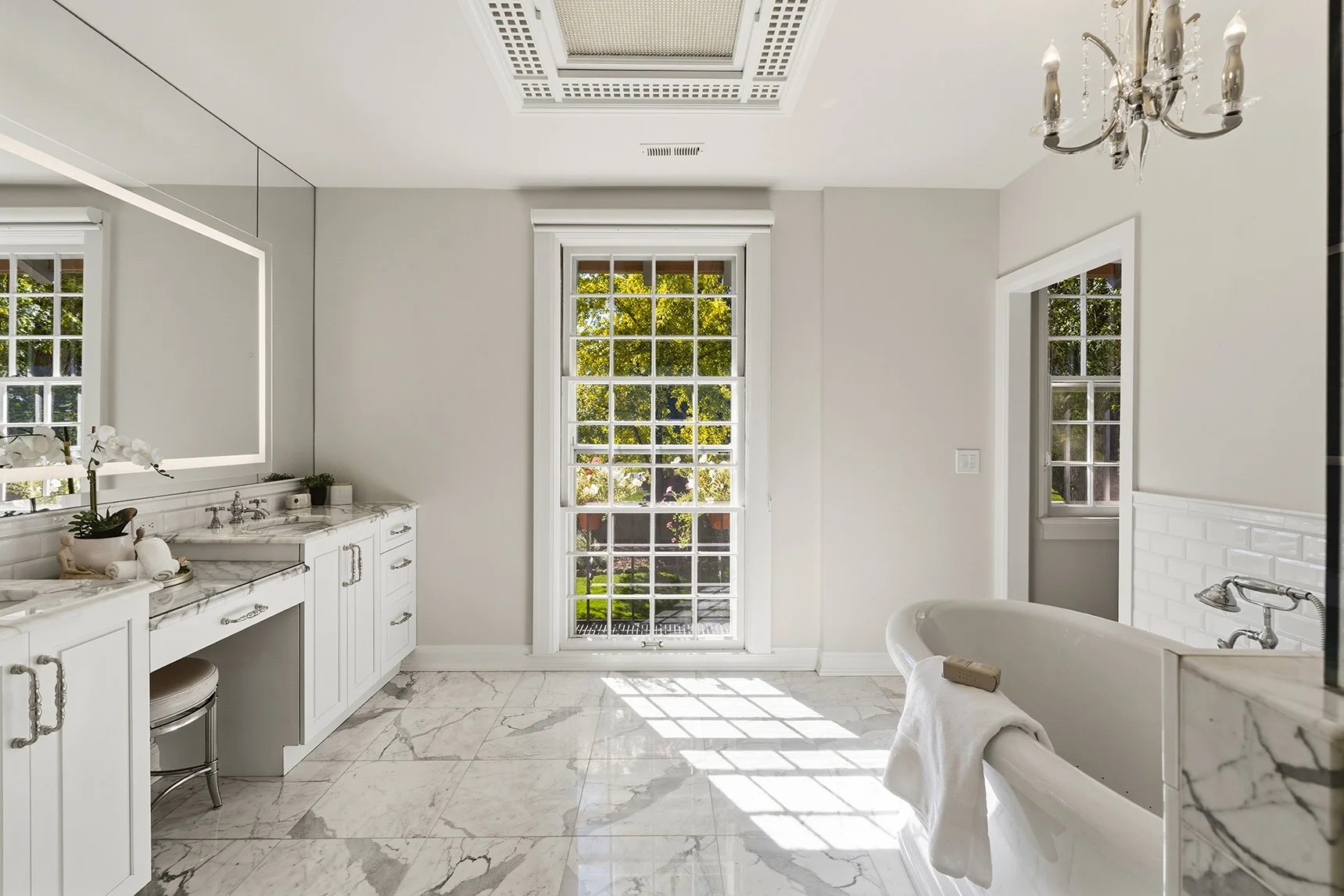 Bright bathroom with a large window showing trees outside, white cabinetry with marble countertops, a bathtub with a towel, and an elegant chandelier.