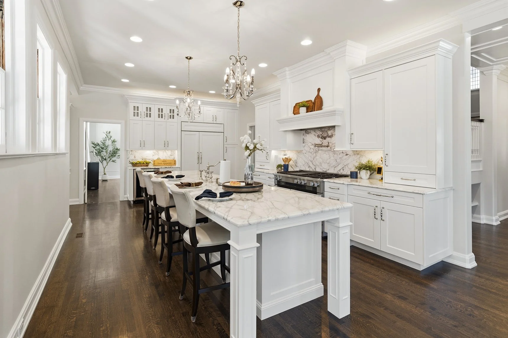 Luxurious white kitchen with marble countertops, large island, pendant chandeliers, custom cabinetry, and dark hardwood floors