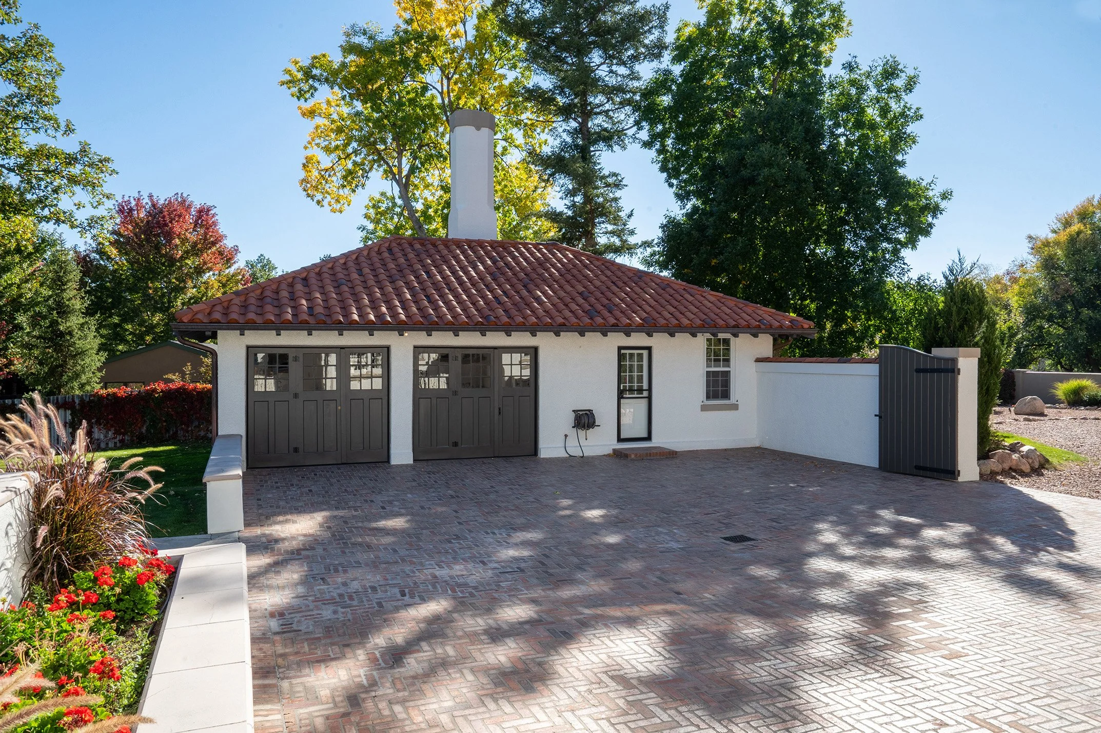 A white house with a red tile roof, two gray garage doors, and a small window. The front yard is paved with bricks and is surrounded by lush trees and colorful flowers.
