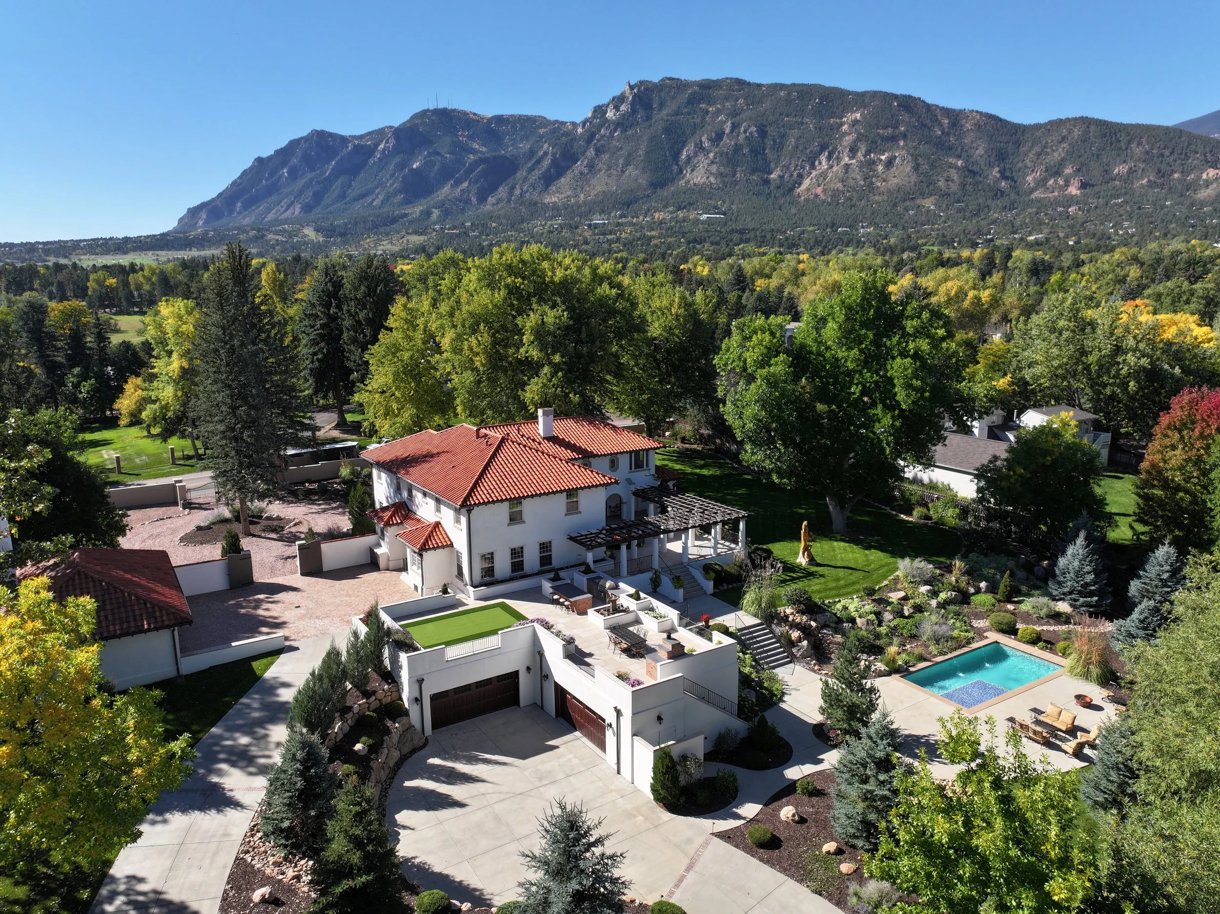 A scenic view of a residential backyard with a swimming pool, surrounded by lush green trees and landscaped gardens, with a mountain range in the background under a clear blue sky.