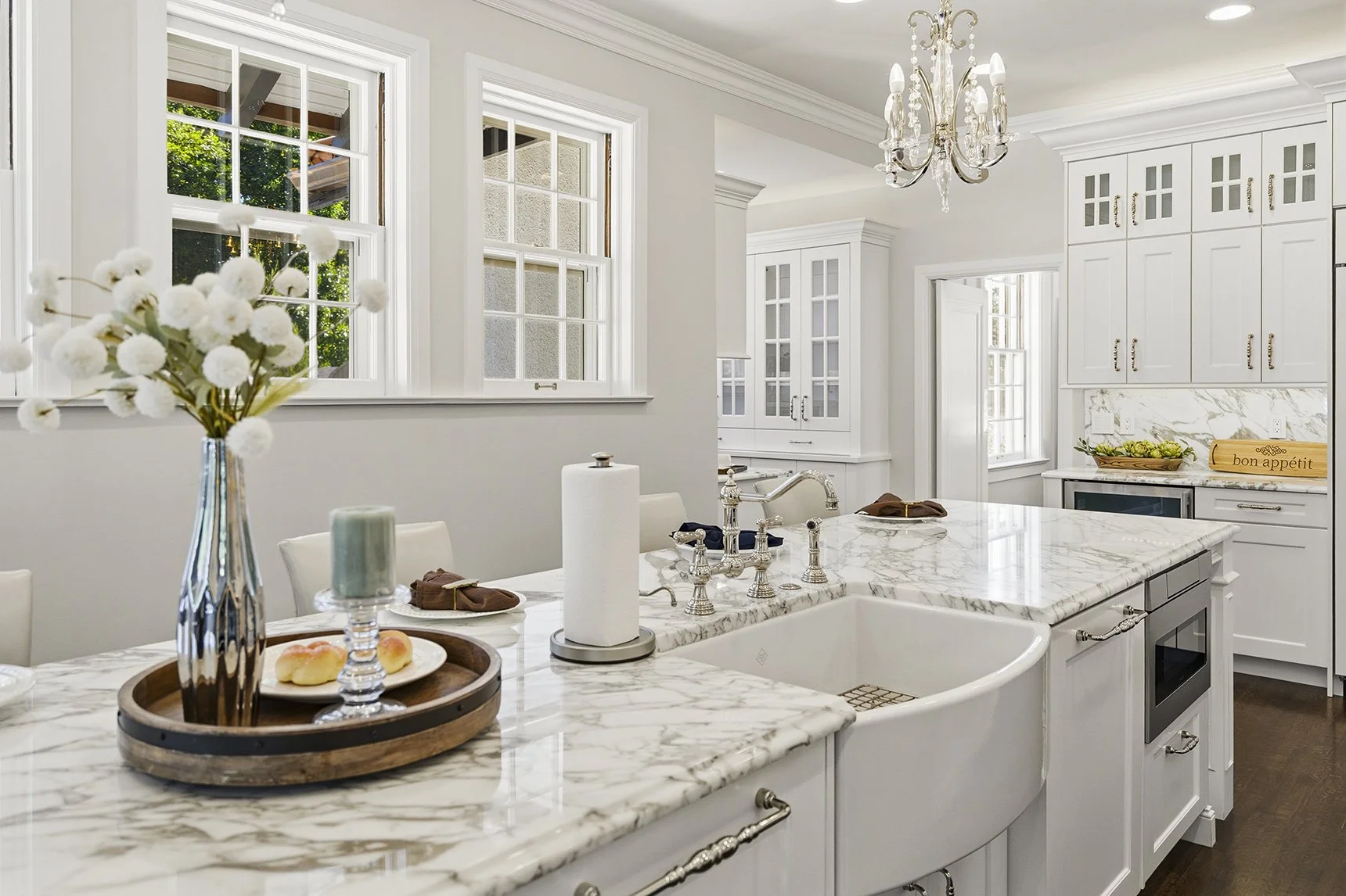 A bright, modern kitchen with white cabinetry, a marble island with a built-in sink, and a chandelier overhead. There are three large windows letting in natural light, a vase with white flowers on the counter, and a loaf of bread with 