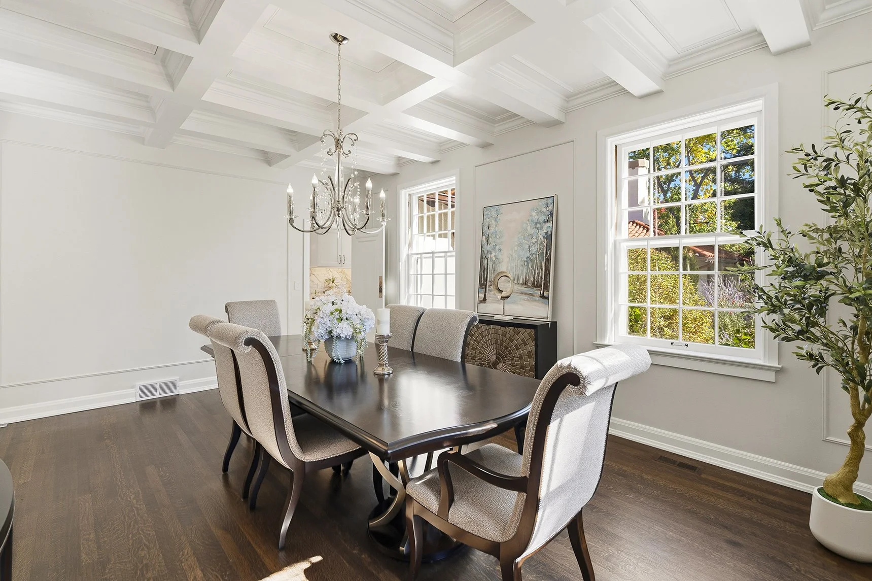 Bright elegant dining room with a dark wood table, upholstered chairs, chandelier, floral centerpiece, coffered ceiling, artwork, and large windows.