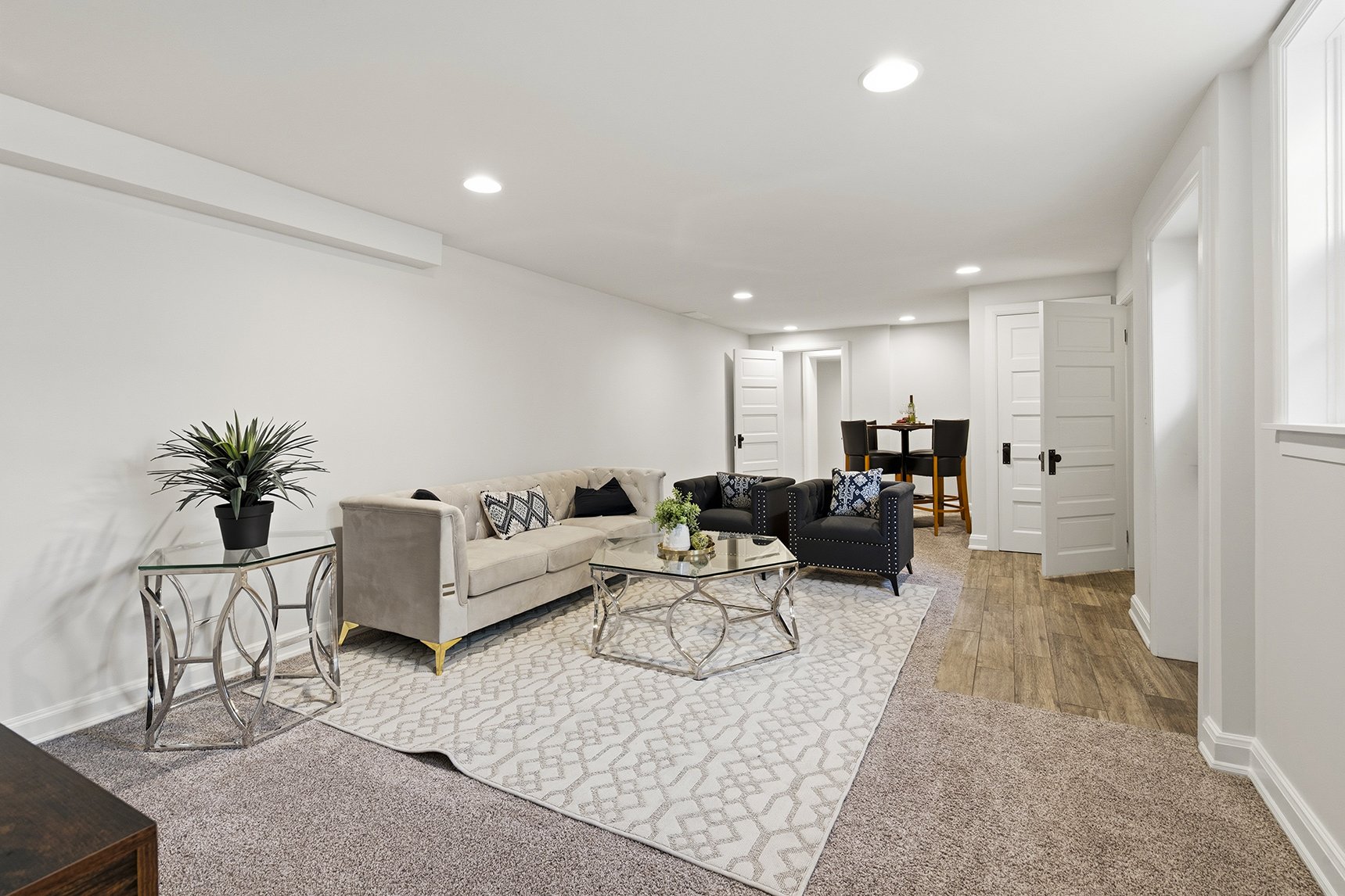Bright finished basement living room with a beige sofa, black accent chairs, glass coffee table, patterned area rug, potted plant, and adjoining dining area