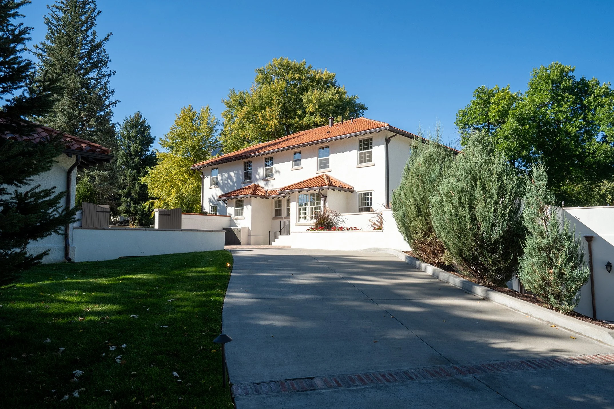 White stucco house with red tile roof, driveway, landscaped yard, and mature trees under a clear blue sky