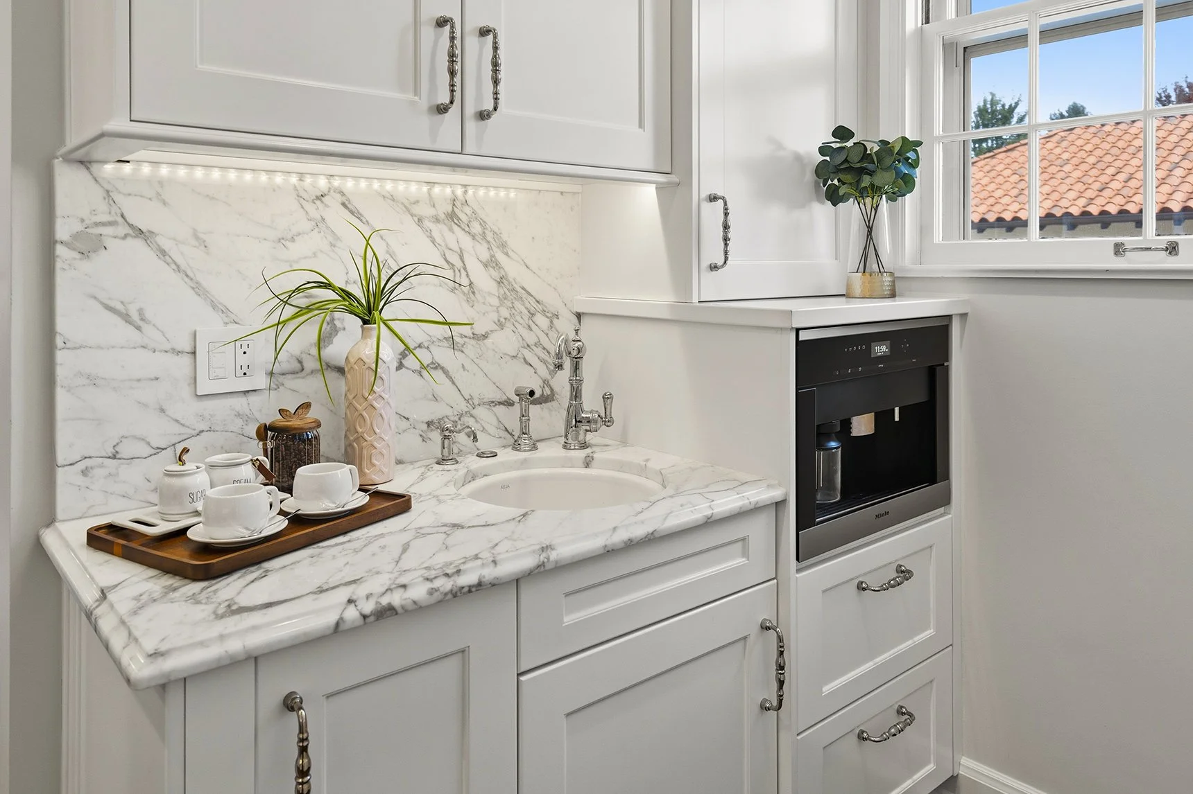 Elegant white butler's pantry with marble countertops, farmhouse sink, coffee maker, cabinetry, and window