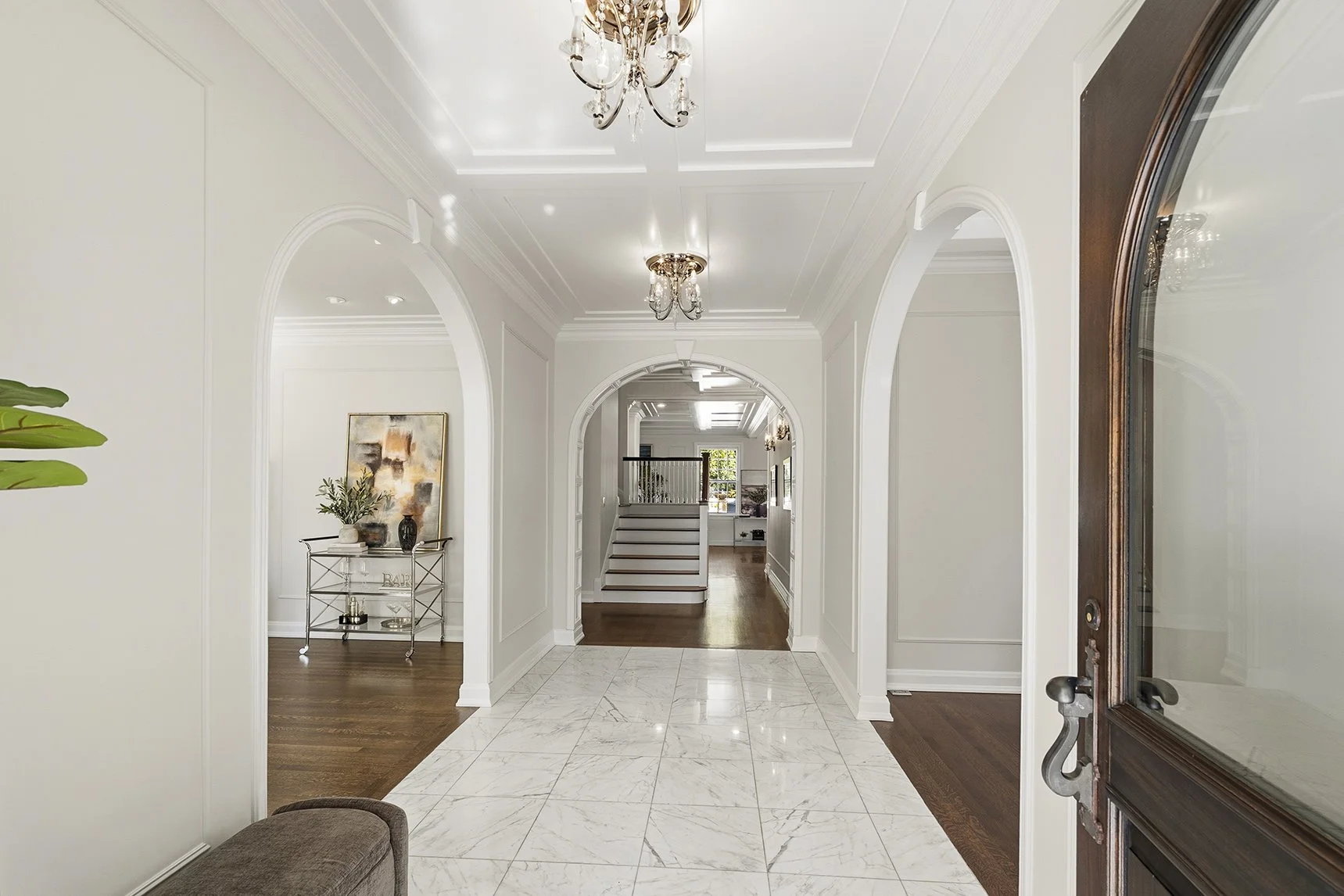 View of a house entrance hall with white walls, marble flooring, arched doorways, a chandelier, and a staircase in the background.