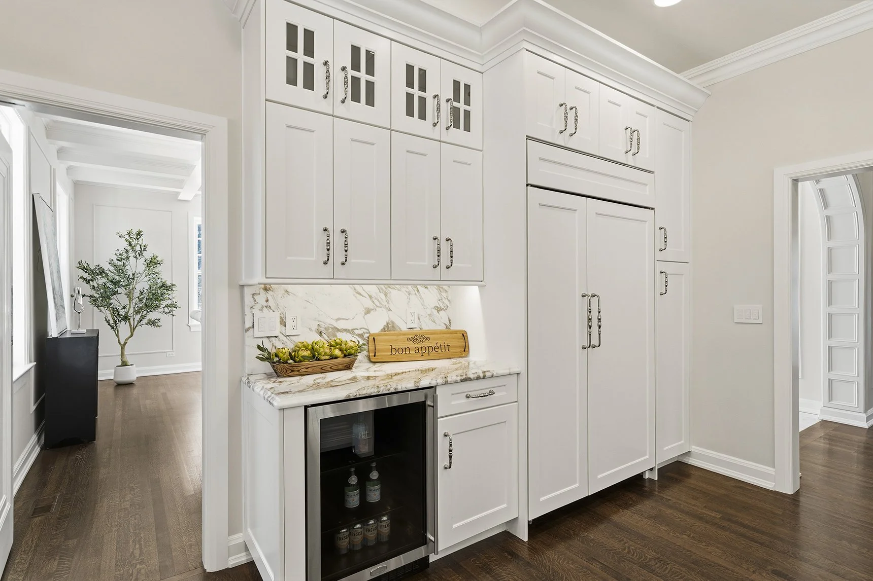 Bright white kitchen pantry with ornate cabinets, marble backsplash and countertop, beverage refrigerator, and dark hardwood floors
