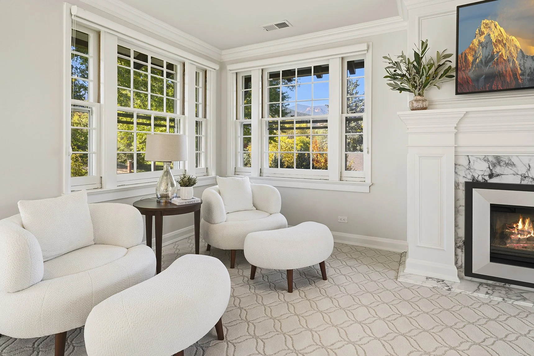 Living room with white sofa and armchair, large windows showing outside trees with fall colors, fireplace with marble surround, mounted flat-screen TV, and a ceiling vent.