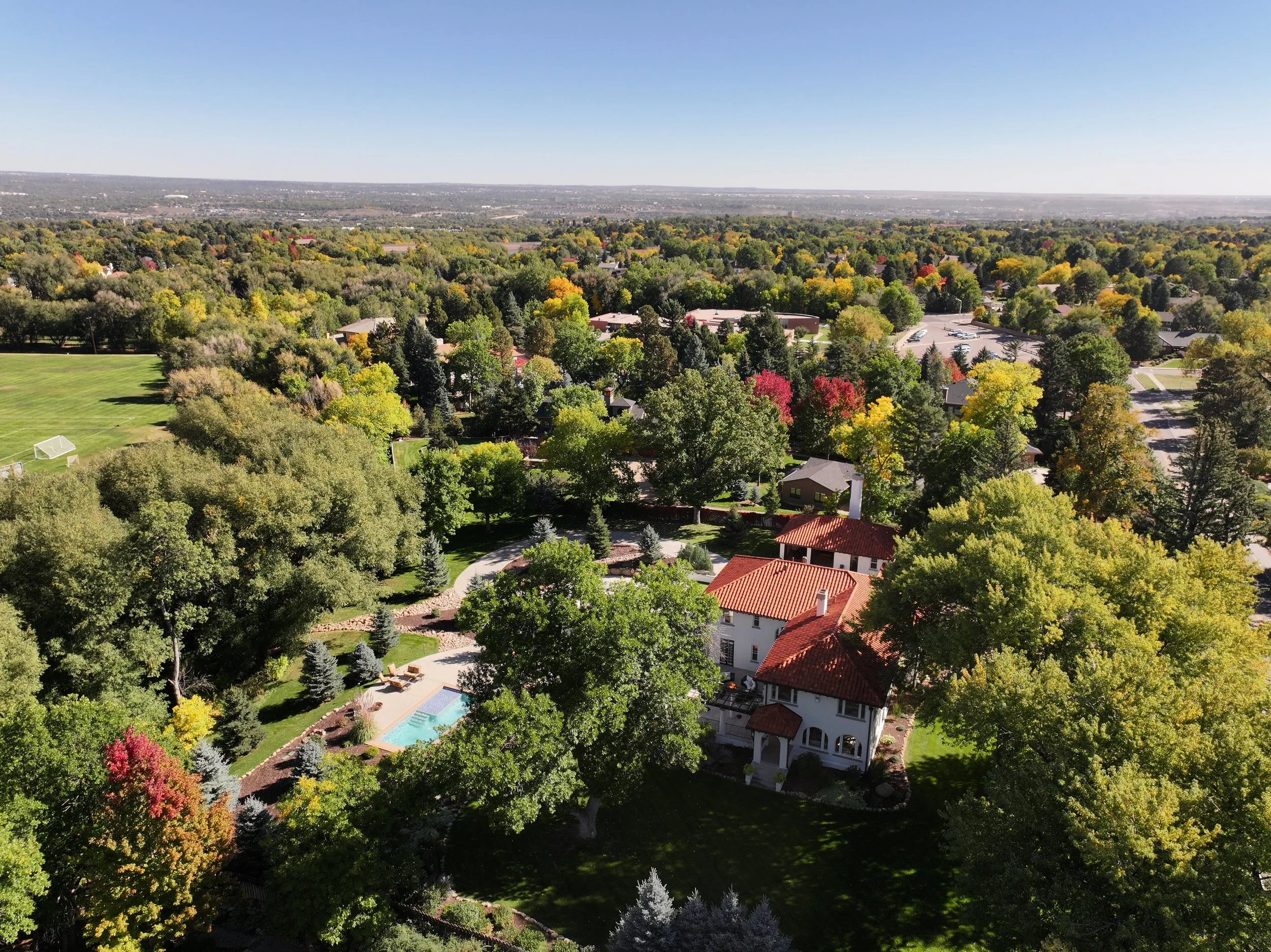 Aerial view of a leafy suburban neighborhood with autumn trees, homes, a swimming pool, soccer field, and distant landscape under a clear blue sky.