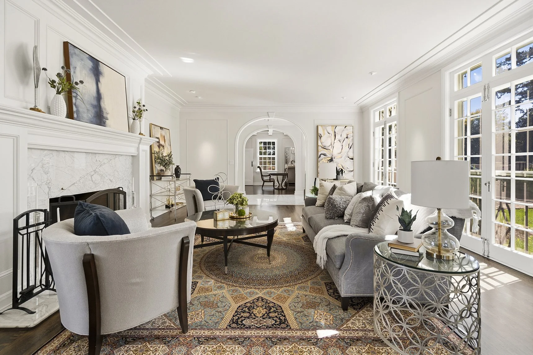 Bright elegant living room with fireplace, gray sofa, armchairs, patterned area rug, and large French doors