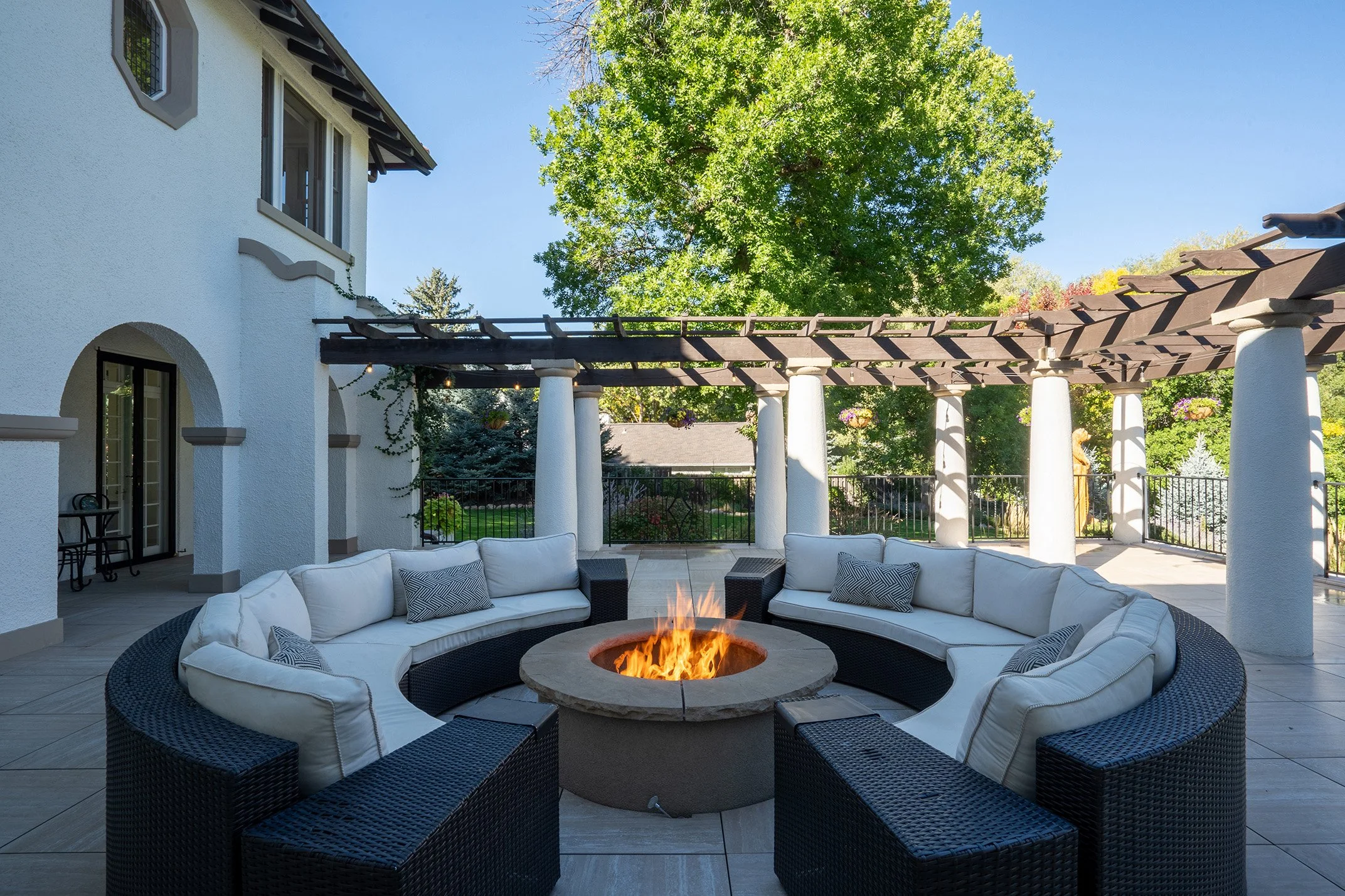 Luxury outdoor patio with a circular fire pit, cushioned wicker seating, and a wood pergola beside a stucco home