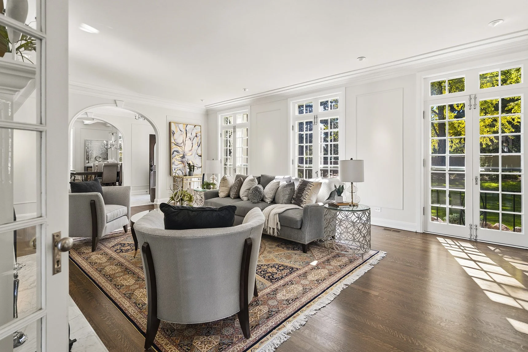 Bright elegant living room with gray sofas and armchairs, patterned area rug, large French doors, and an adjoining dining room.