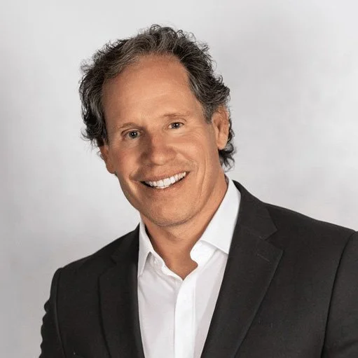 A smiling man with curly almost balding hair in a black suit and white shirt, against a light background.