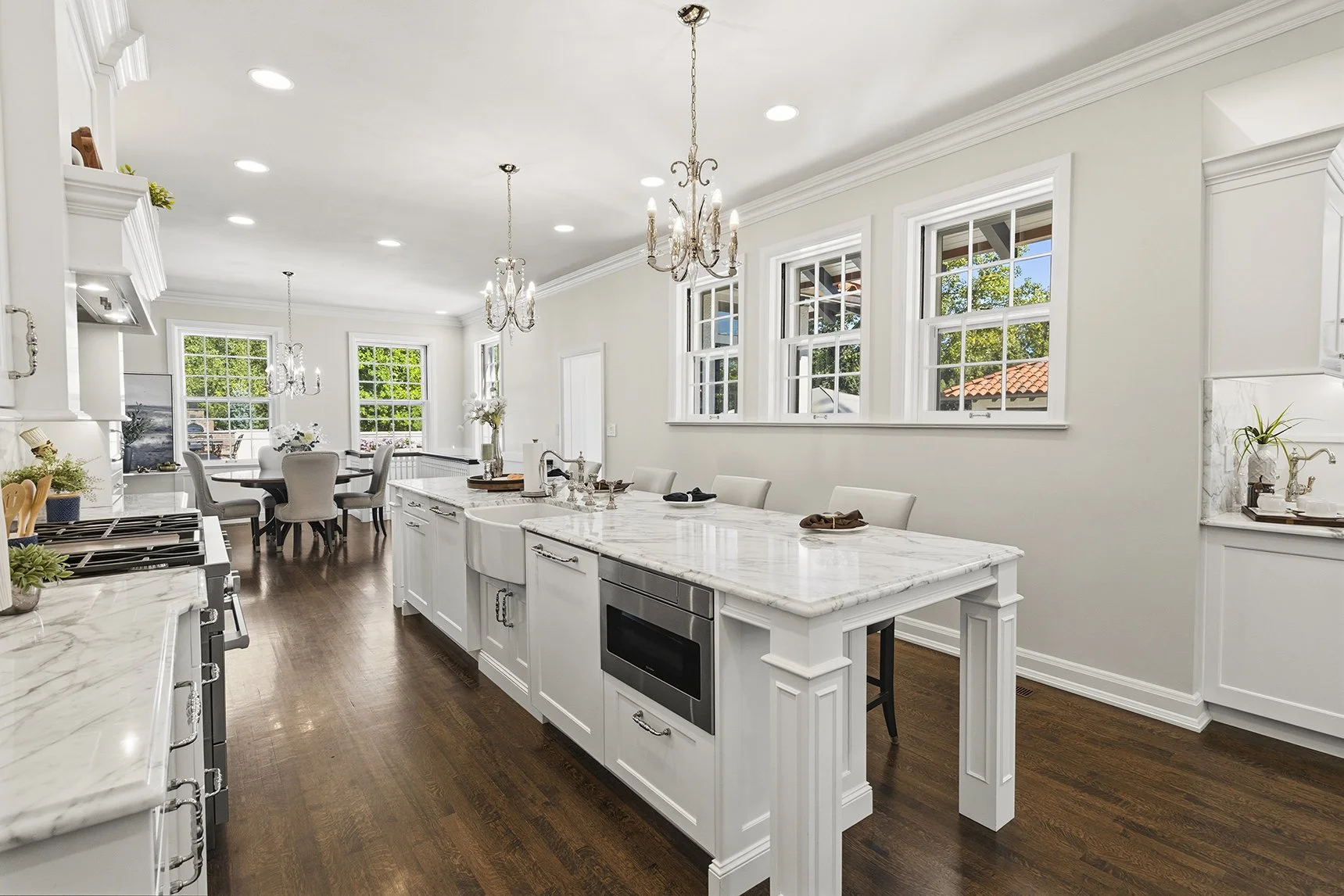 Bright luxury white kitchen with marble countertops, large island, chandeliers, stainless steel appliances, and adjacent dining area