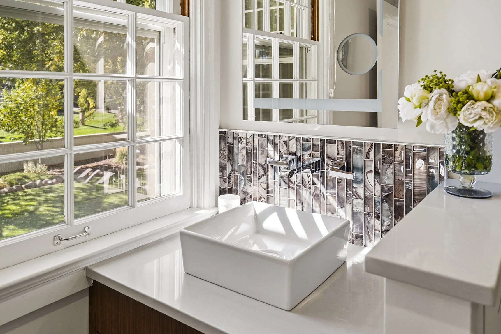Bright modern bathroom with large windows, white vessel sink, geometric tile backsplash, round mirror, and floral arrangement