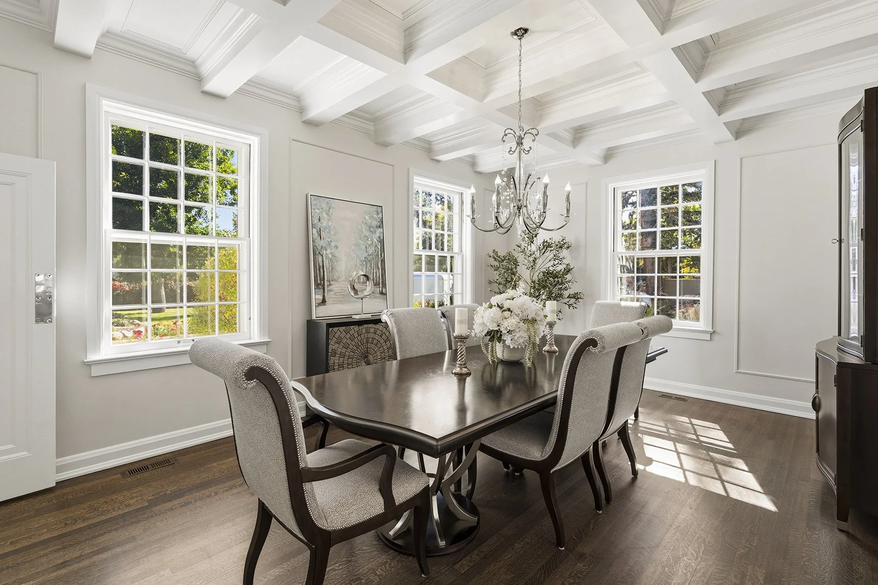 A bright dining room with white walls, large windows, and a white coffered ceiling. There is a dark wood dining table with six upholstered chairs, a chandelier hanging above, and a floral centerpiece. The room has hardwood floors and a modern paintin