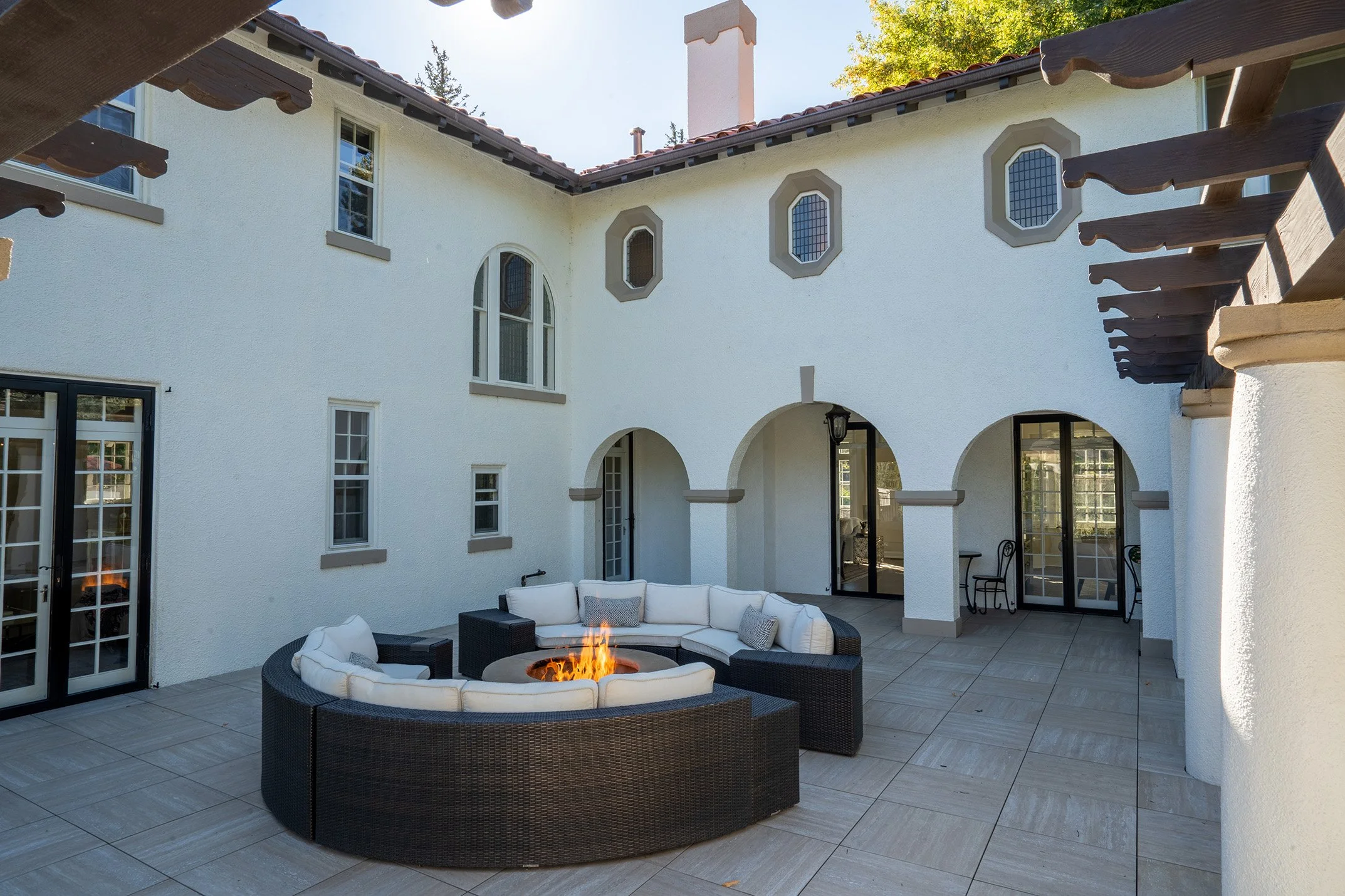 Spanish-style courtyard patio with arched doorways, pergola, tiled roof, and circular wicker seating around a fire pit