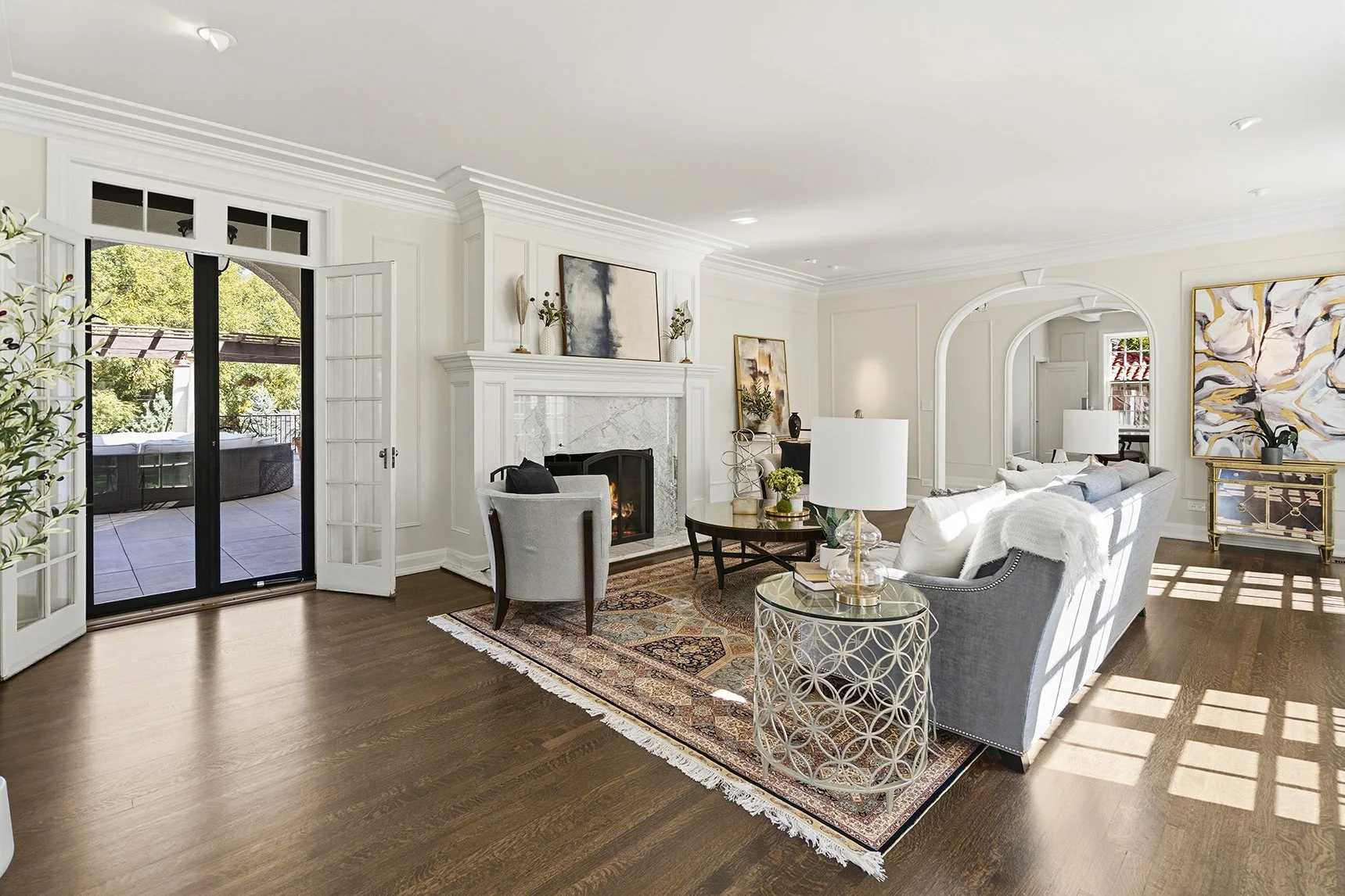 Bright, elegant living room with a marble fireplace, gray sofas, patterned rug, white furnishings, and French doors opening to a patio.