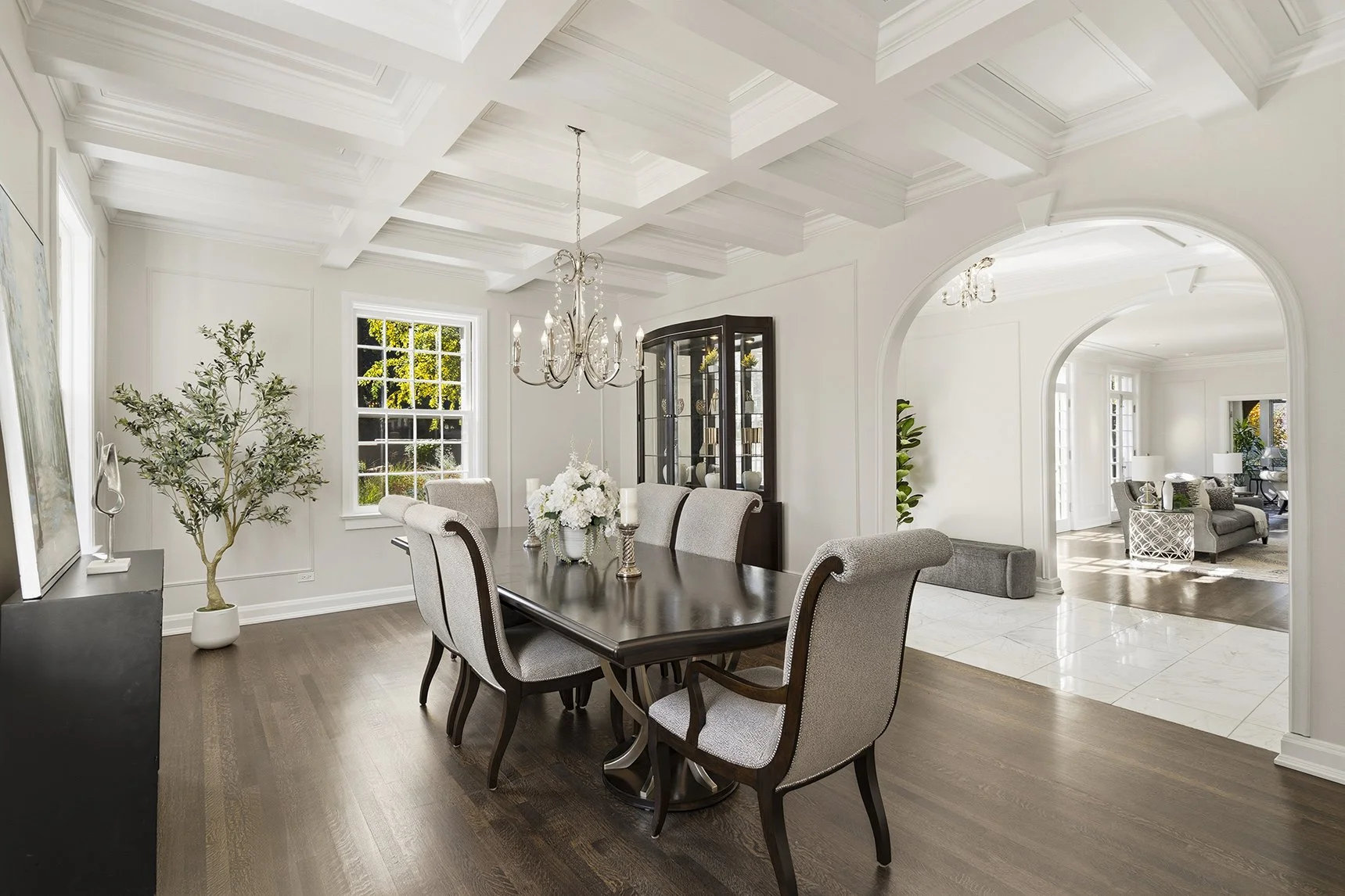 Dining room with white walls, wooden floor, dark wood table, upholstered chairs, chandelier, and large windows showing outdoor greenery.