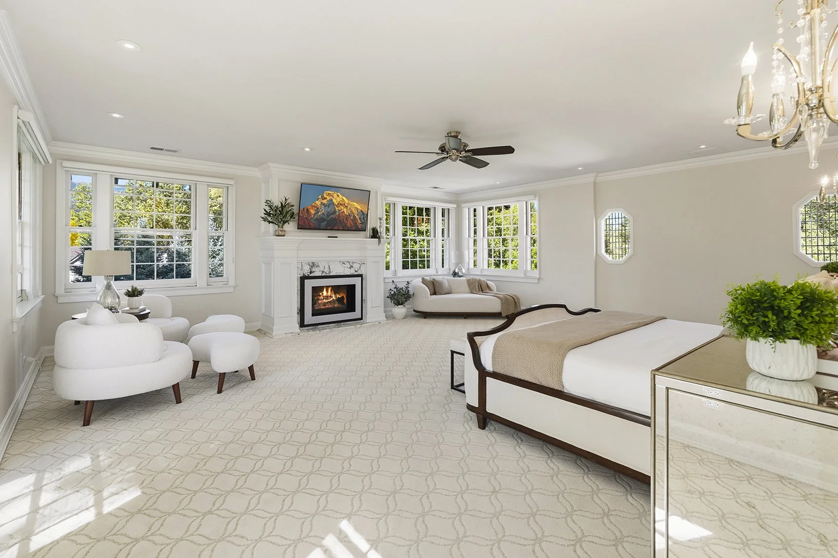 Bright and airy living room with white walls, large windows, a fireplace, a flat-screen TV, beige and white furniture, a patterned carpet, and decorative plants.