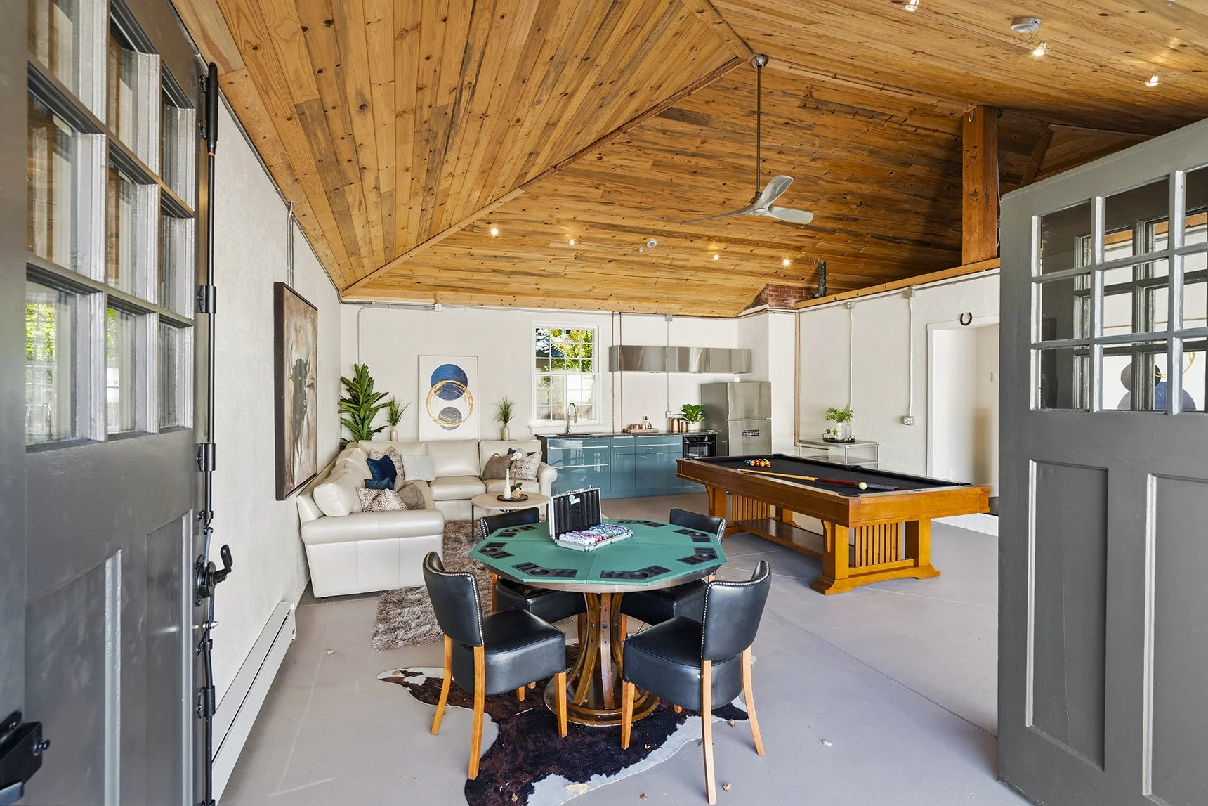 Rustic open-concept game room with a wood-planked ceiling, pool table, poker table, sofa, and modern kitchen.